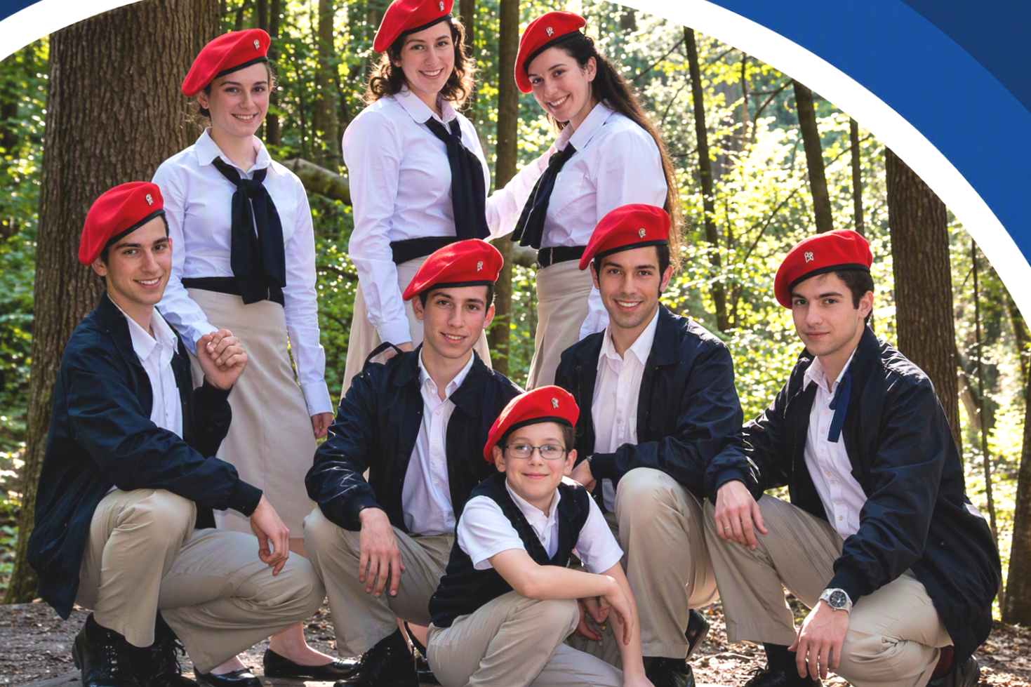 A group of nine young people, wearing a uniform of white shirts, beige pants, and red berets, posing together in a forested area during daytime.