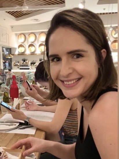 A young woman with brown hair smiling at the camera while sitting at a restaurant table with a smartphone in her hand.