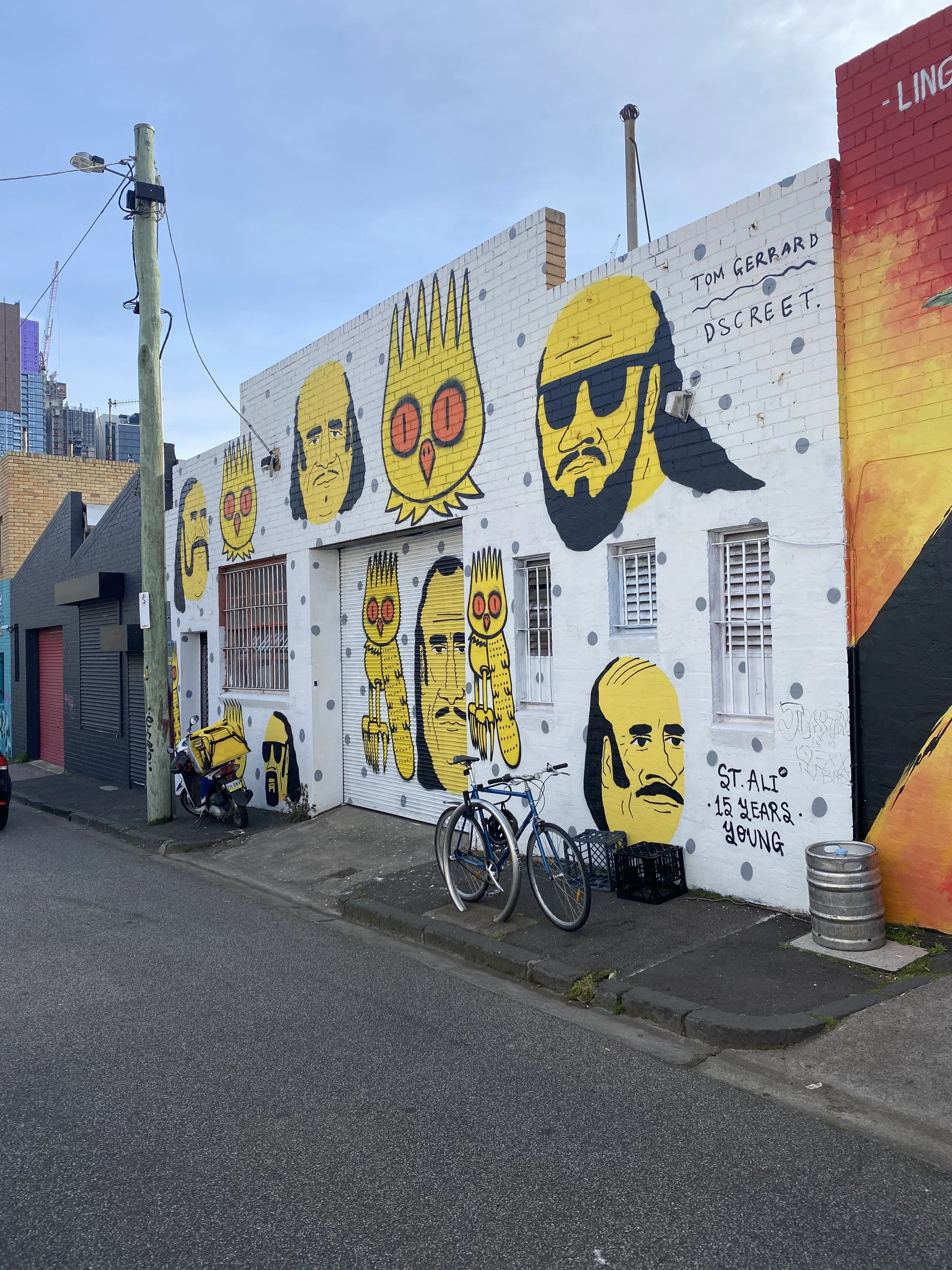 Street view of a building with colorful street art murals, including various cartoon-like human and animal faces. There are two bicycles parked on the sidewalk, a motorcycle, and a metal trash bin. The murals are painted in yellow, black, red, and white, with some text written in black, including "ST. ALI 15 YEARS YOUNG" and "TOM GERRARD DISCREET." The street is wet, and a utility pole with wires is visible in the foreground.