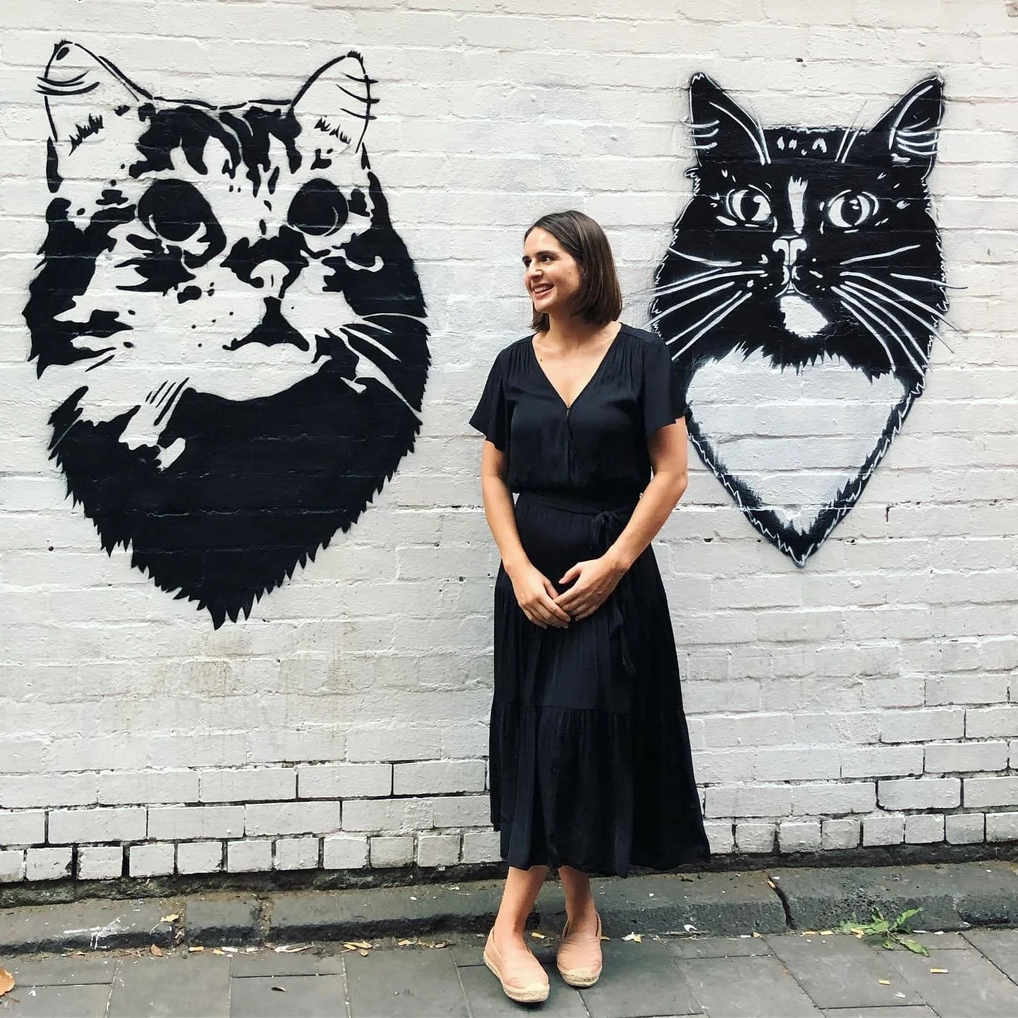 A woman in a black dress standing against a white brick wall with black and white cat head murals on each side.