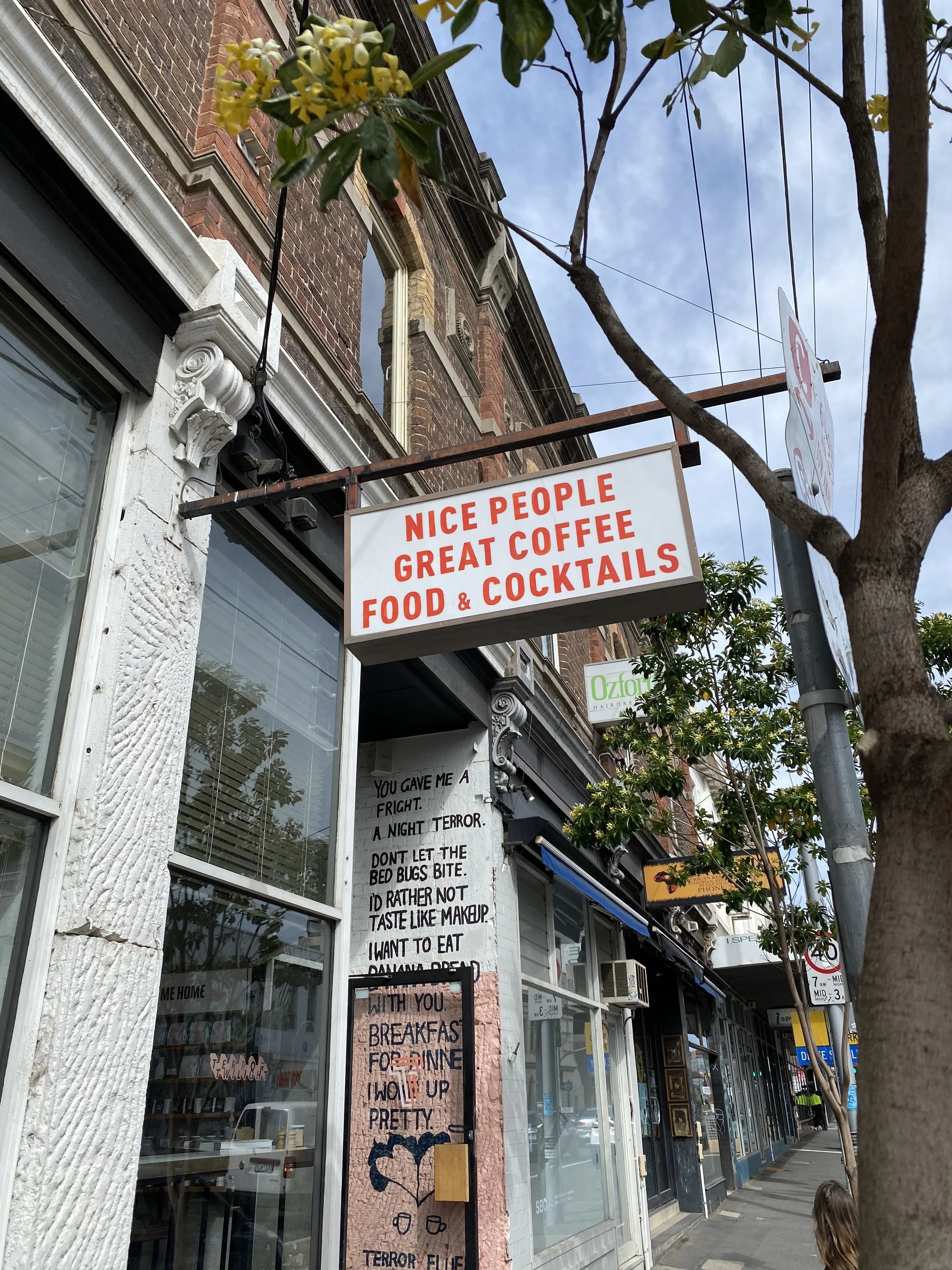 A street view showing a building with a sign advertising a cafe or bar that offers food, coffee, and cocktails. The building has large windows, brickwork, and decorative architectural details. There are also handwritten signs and a tree lining the sidewalk.