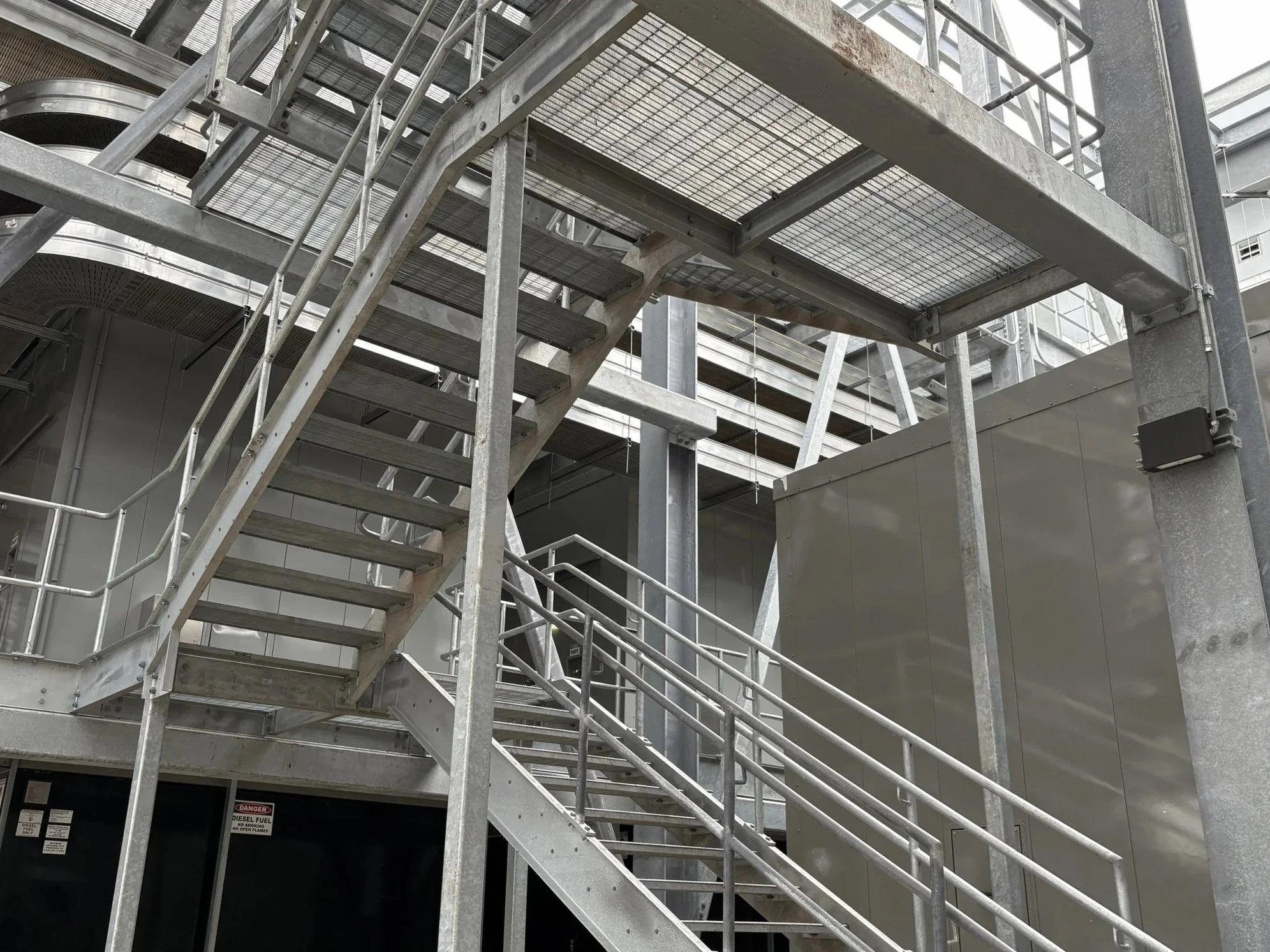 Metal emergency stairway outside a modern building with a gray exterior, metal railings, and a warning sign near the door.