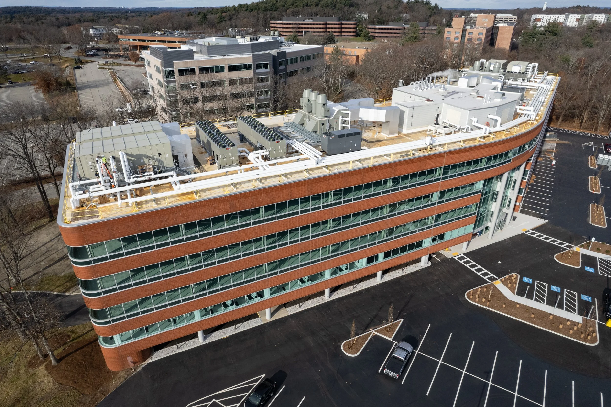 An aerial view of a multi-story commercial building with a flat roof, featuring HVAC systems, ducts, and vents. The building has curved red brick walls and large glass windows, surrounded by parking lots with some parked cars and landscaped areas with trees.