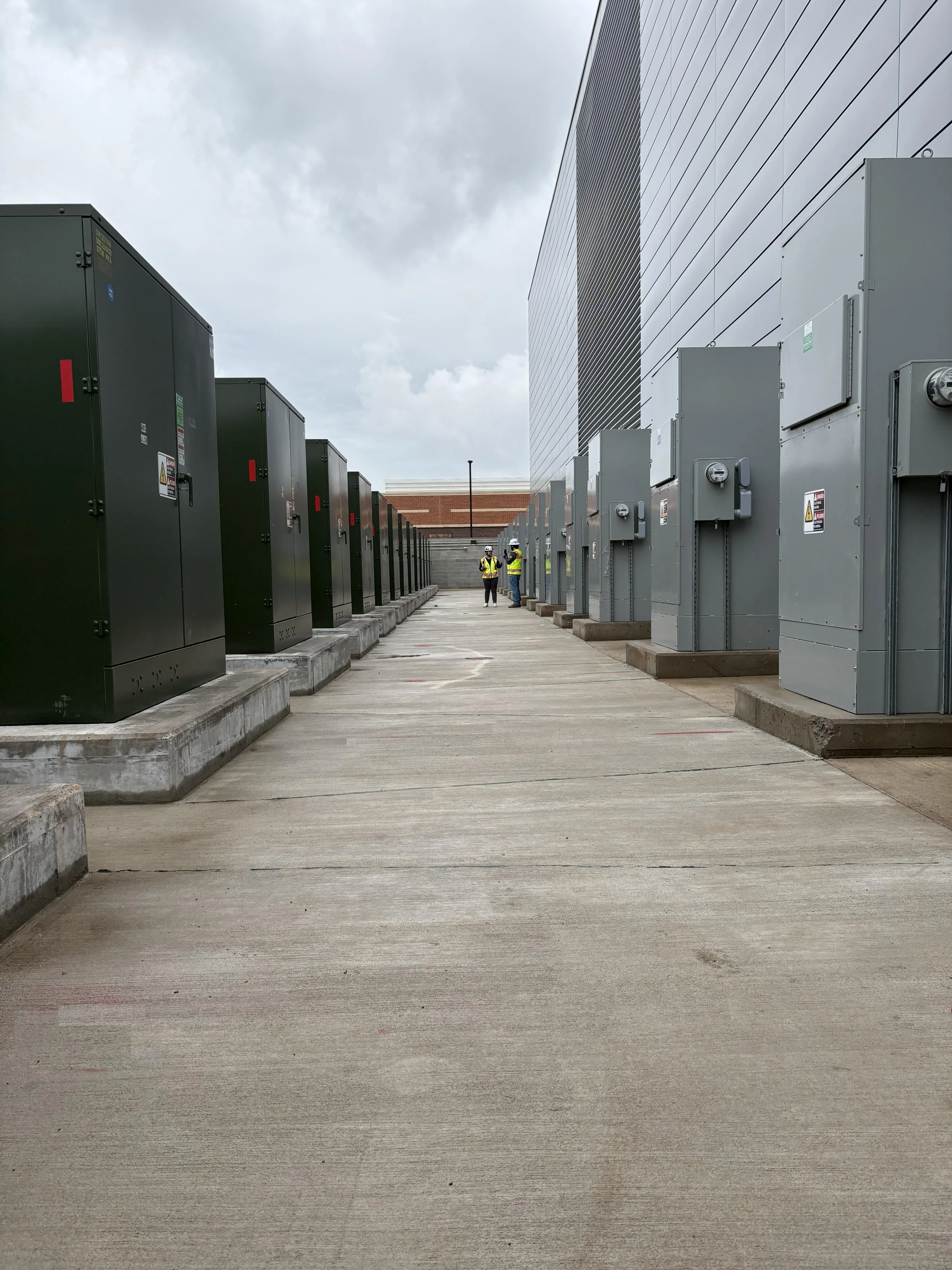 Two workers wearing safety vests and helmets standing in an outdoor area with multiple electrical transformer boxes along a concrete path next to a large modern building, under a cloudy sky.