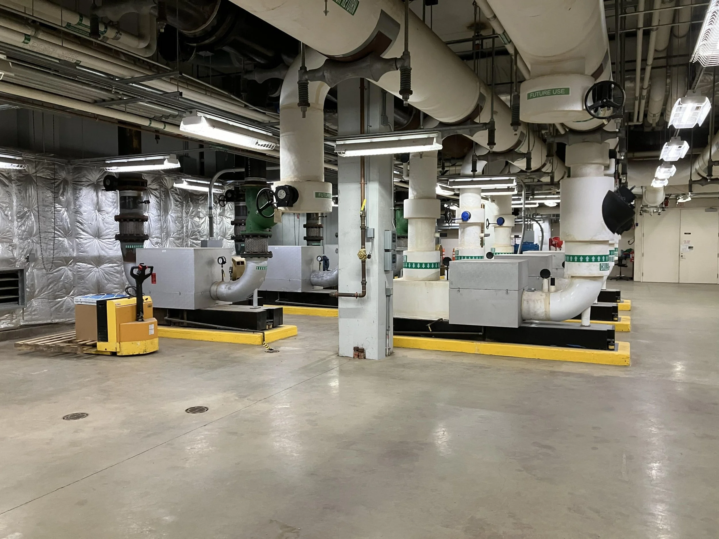 Industrial or mechanical room with large pipes, valves, and equipment, some labeled 'Future Use', with a concrete floor and fluorescent lighting.