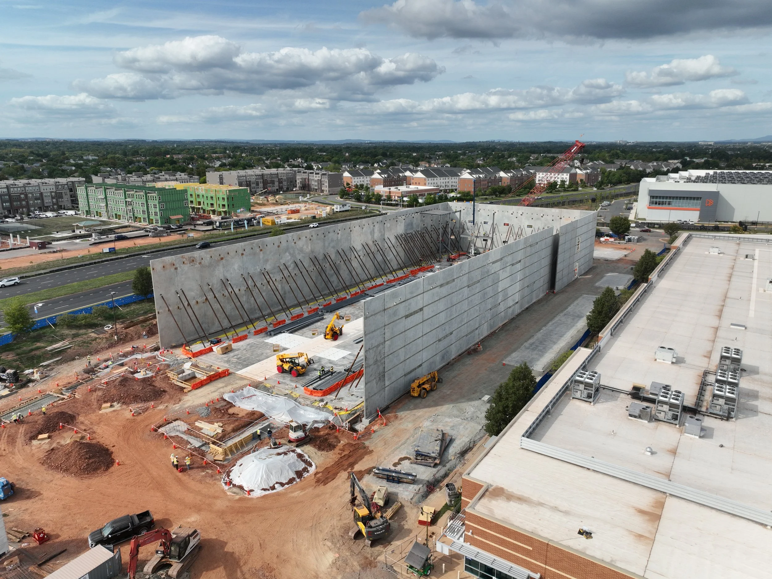 Aerial view of a construction site with a large concrete wall being built, cranes, construction vehicles, and workers, surrounded by residential buildings and a parking lot, under partly cloudy skies.