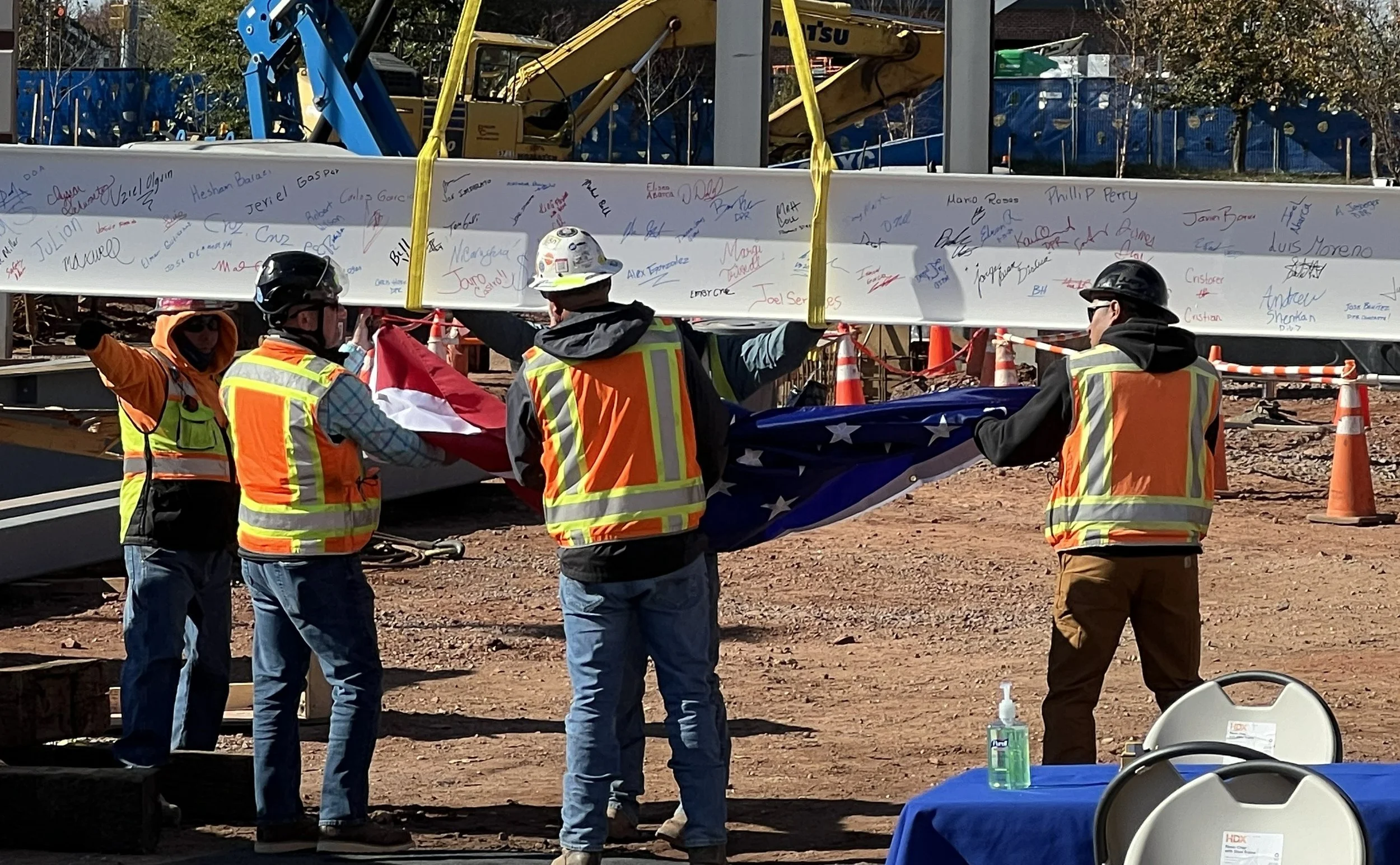 Construction workers at a building site holding a folded American flag, with a large signed beam in the background.