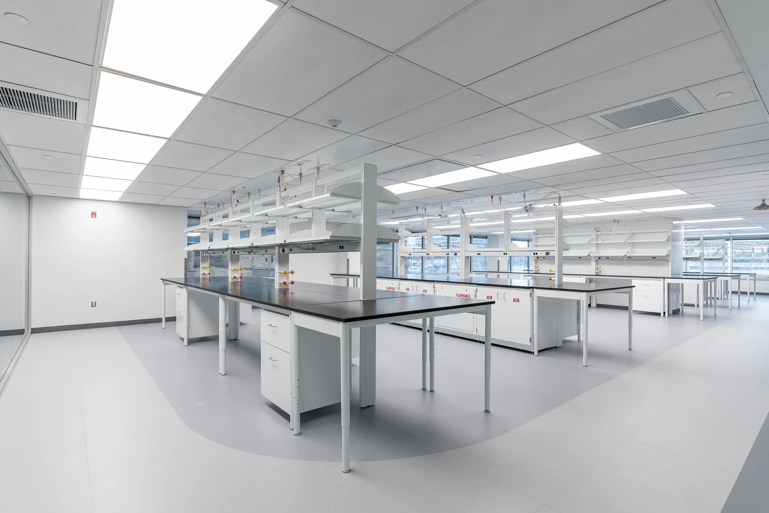 A modern laboratory with white walls, ceiling tiles, and multiple workbenches with black countertops. The lab is equipped with glassware and lab equipment, with large windows providing natural light.