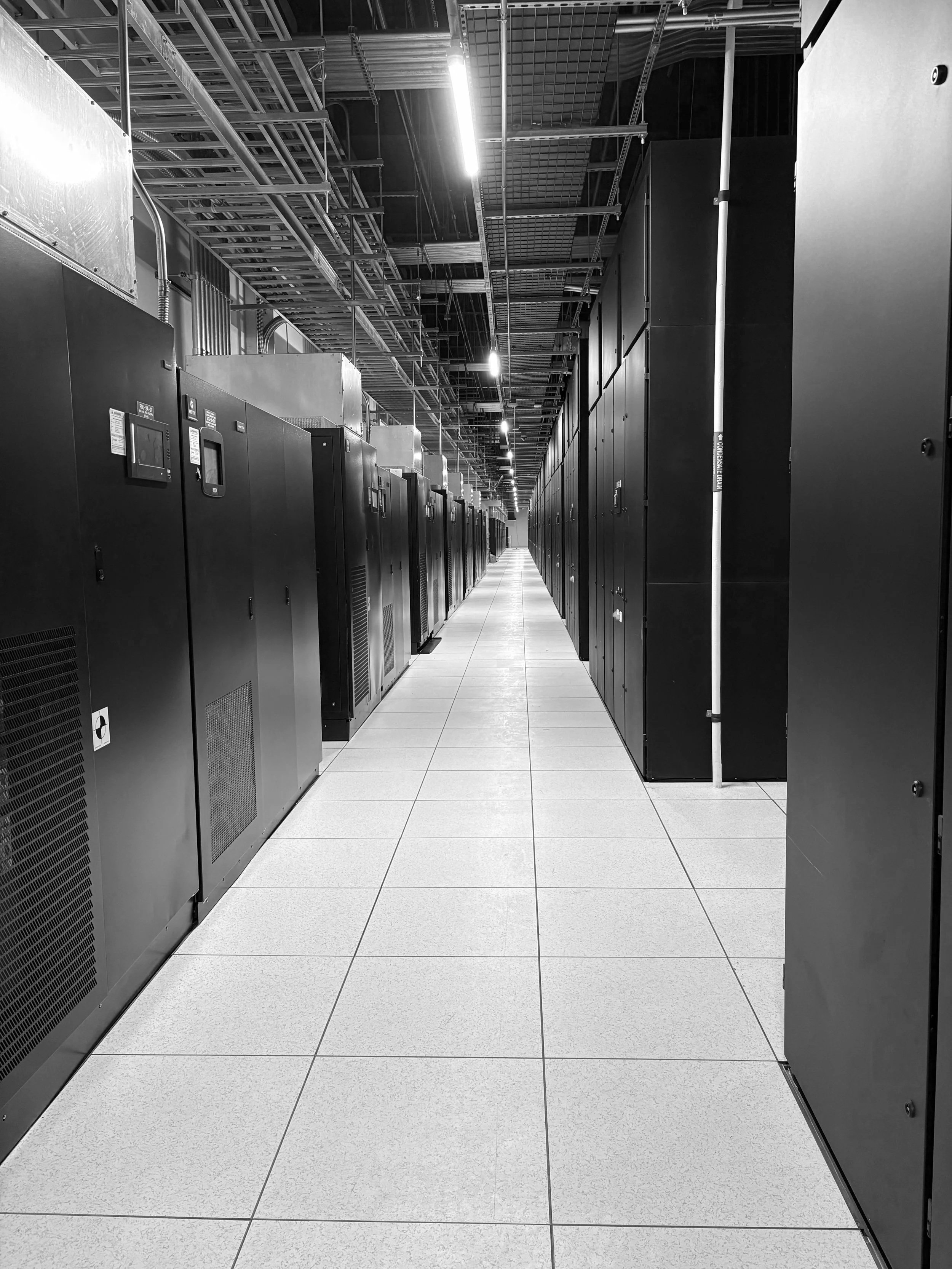 A long aisle in a data center with rows of black server racks on either side, tiled floor, and overhead lighting.