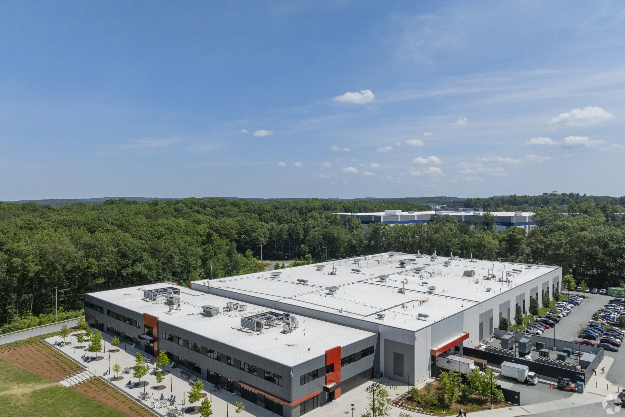 A modern commercial building with a flat white roof, surrounding parking lot filled with cars, and green trees in the background beneath a blue sky with scattered clouds.