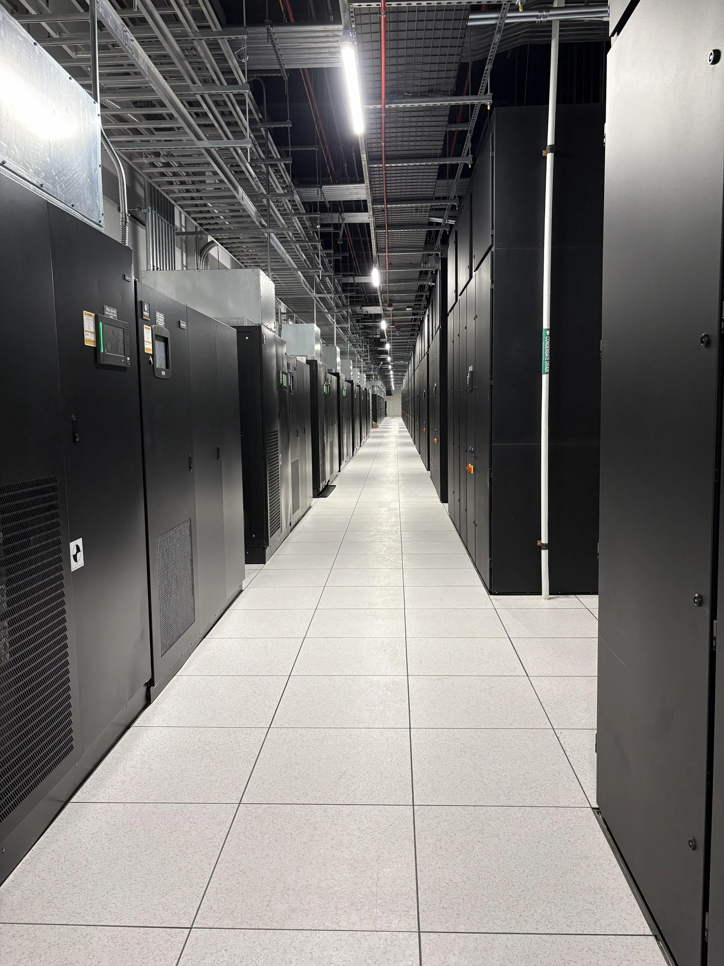 Data center aisle with black server racks on both sides, white tiled floor, overhead cabling, and bright ceiling lights.