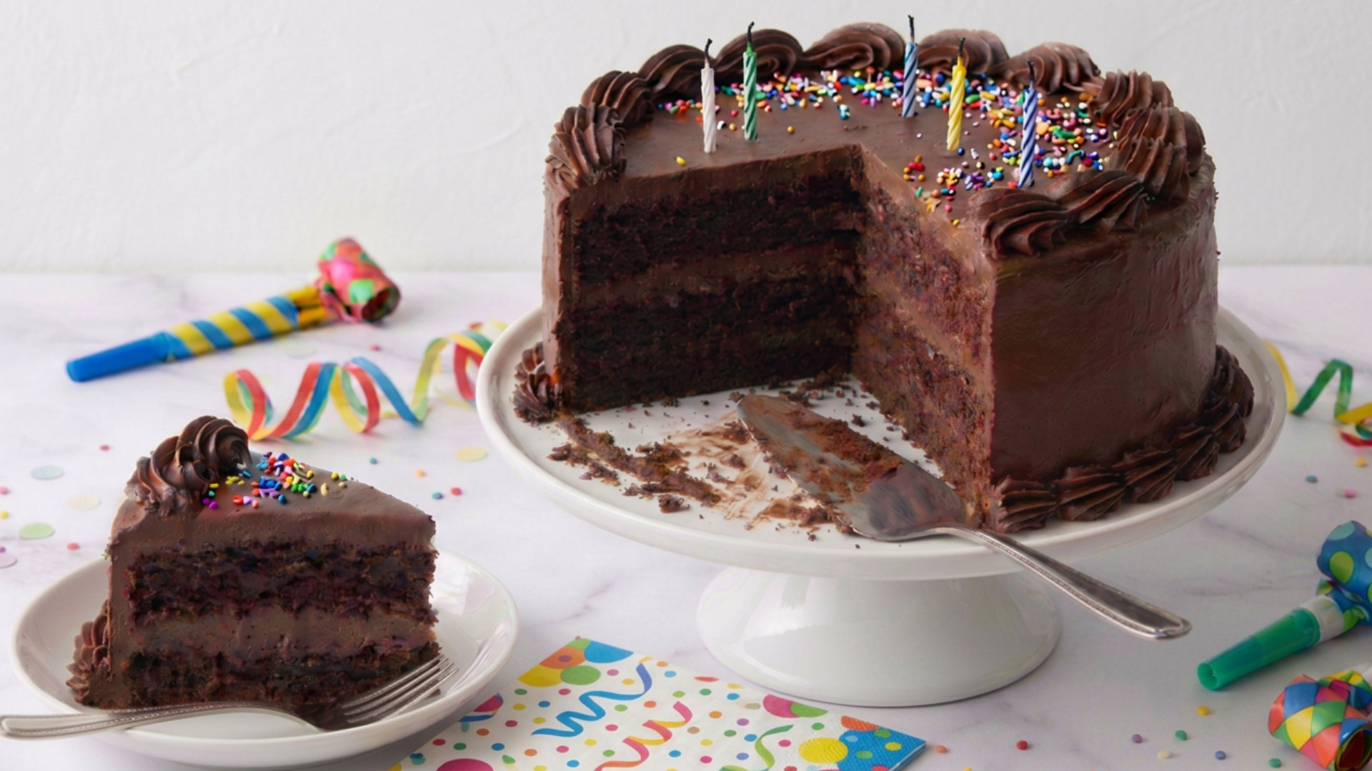Chocolate cake with a side-angle view, full chocolate frosting, chocolate piping on edges, and sprinkles on white cake stand surrounded by party decor