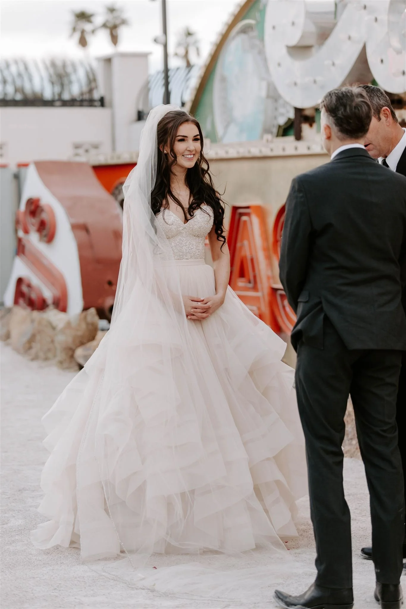 A bride in a white wedding gown and veil smiling at two men in suits during a wedding ceremony outdoors.