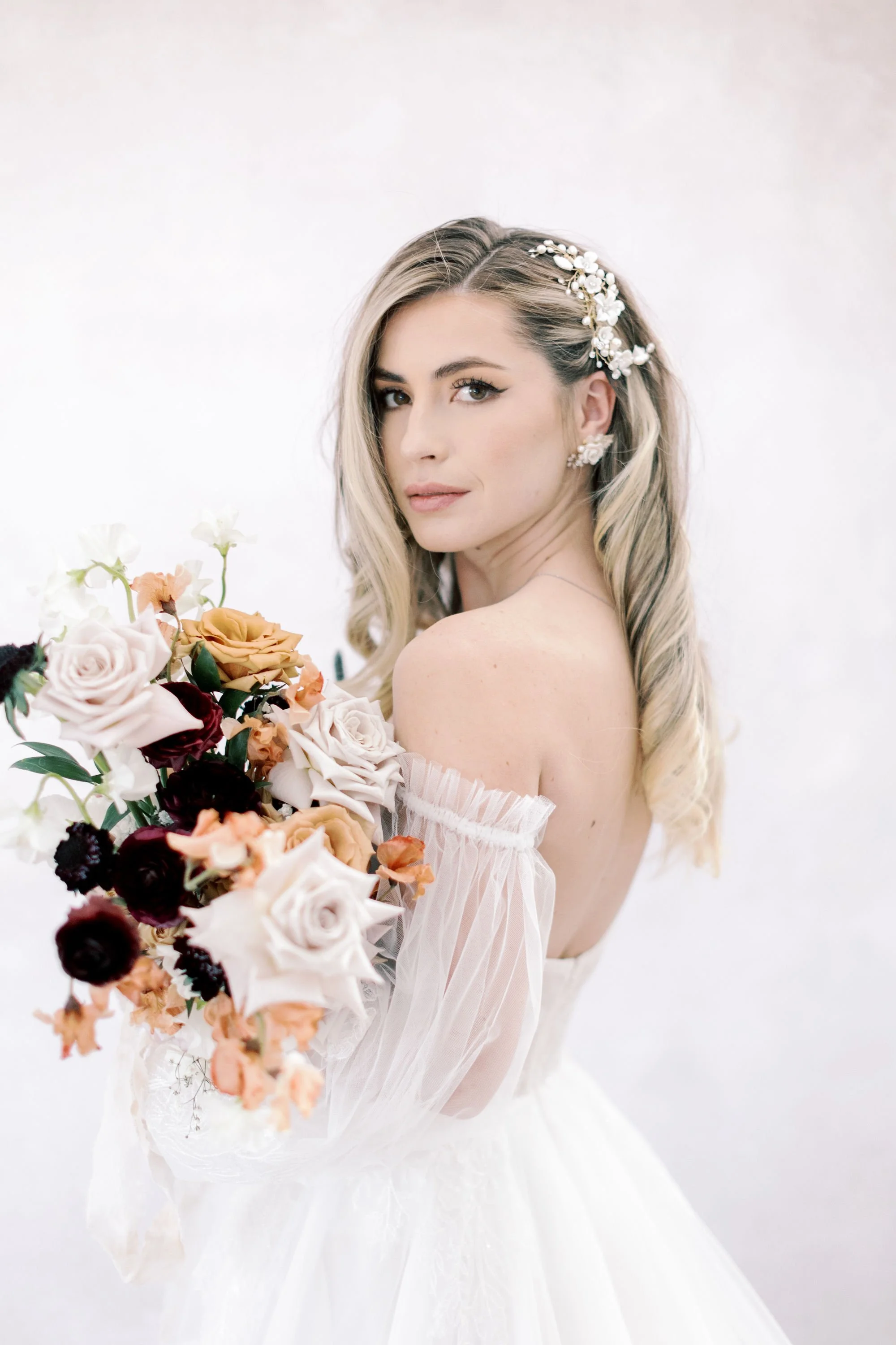 A woman with long, blond, wavy hair adorned with a pearl and floral hairpiece, wearing earrings, and holding a bouquet of multicolored roses, dressed in a white strapless dress, against a plain white background.