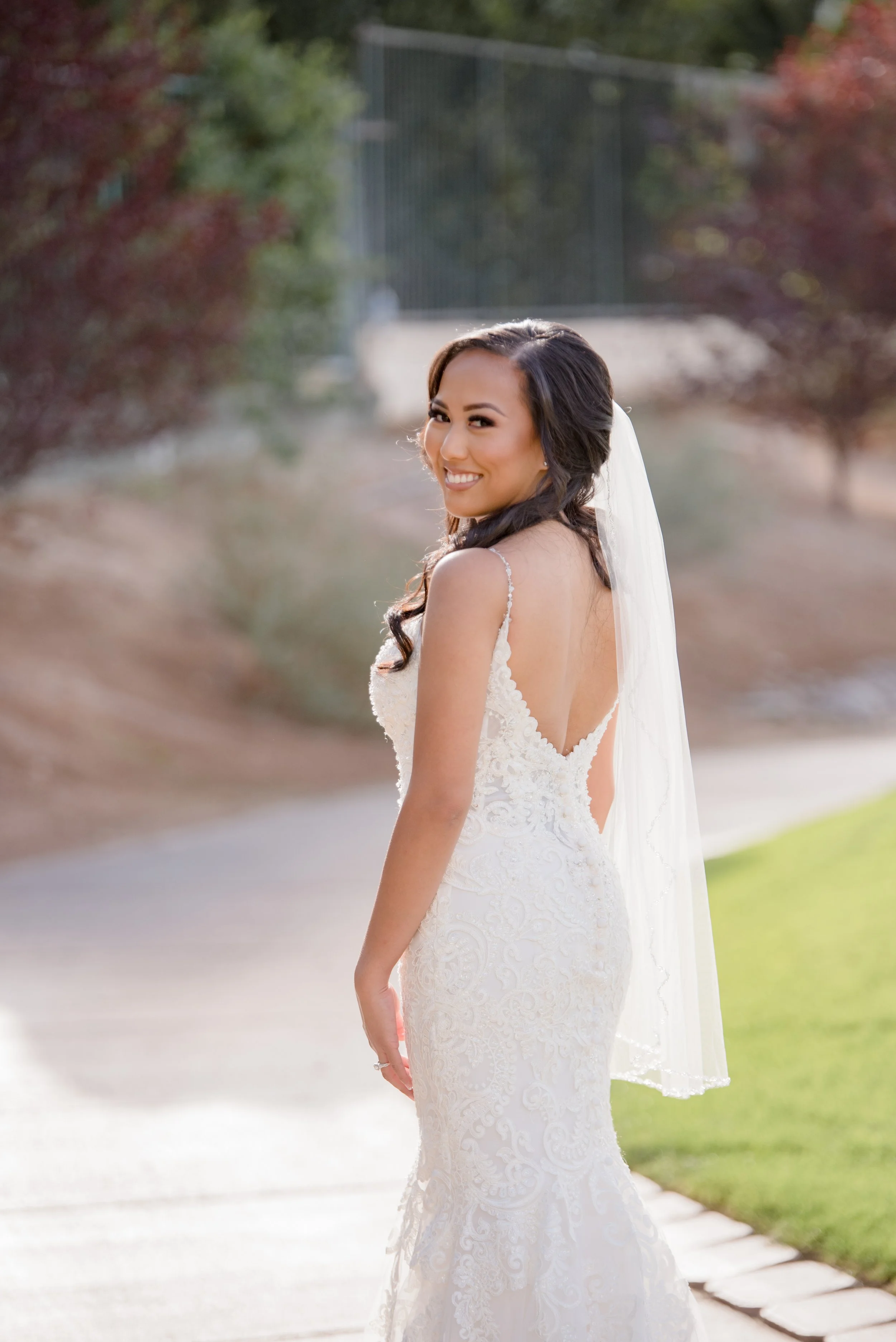 A smiling bride in a white lace wedding gown with a veil, standing outdoors on a sidewalk with trees and greenery in the background.