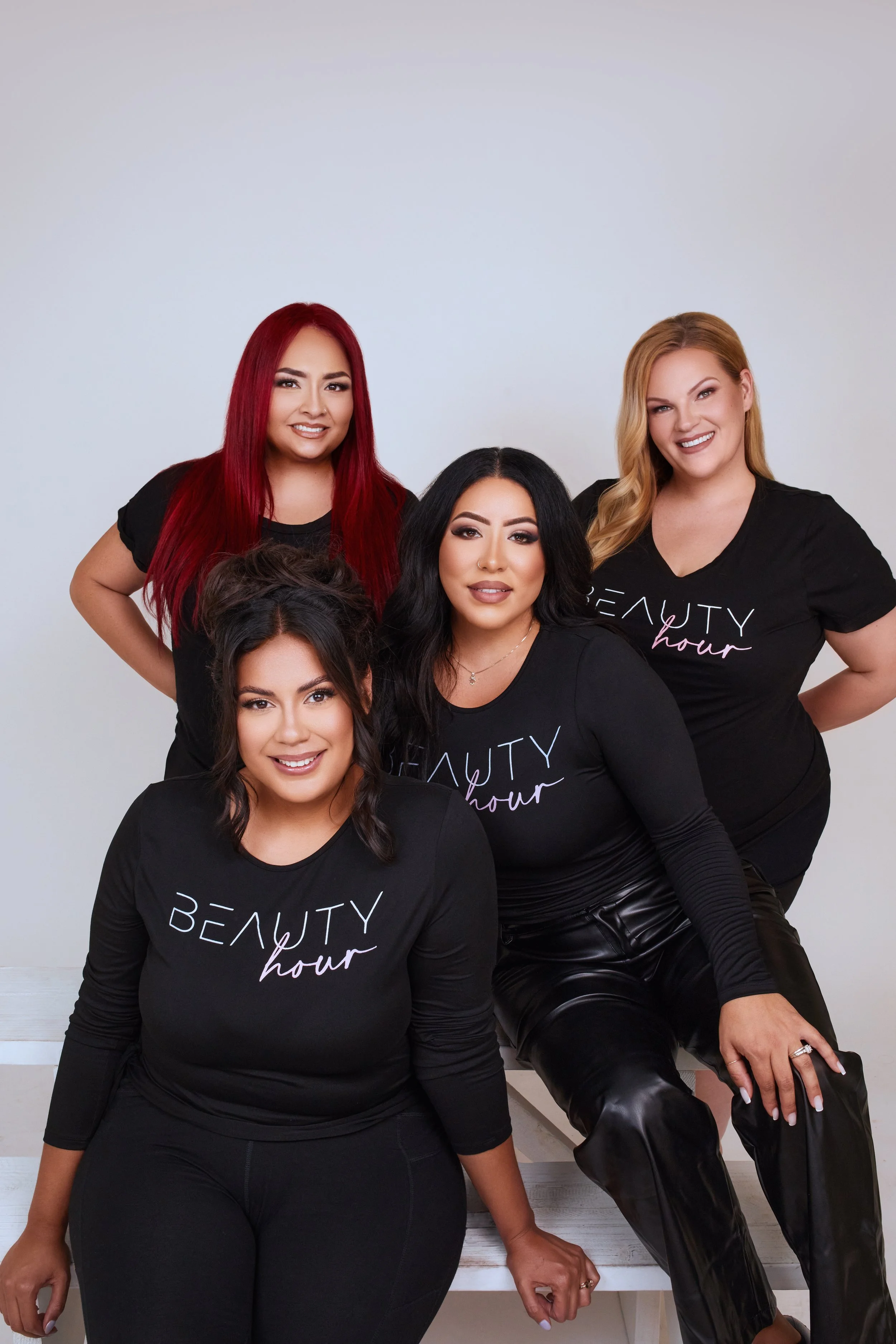 Group of five women posing together, wearing black shirts with 'BEAUTY hour' written on them, smiling at the camera.