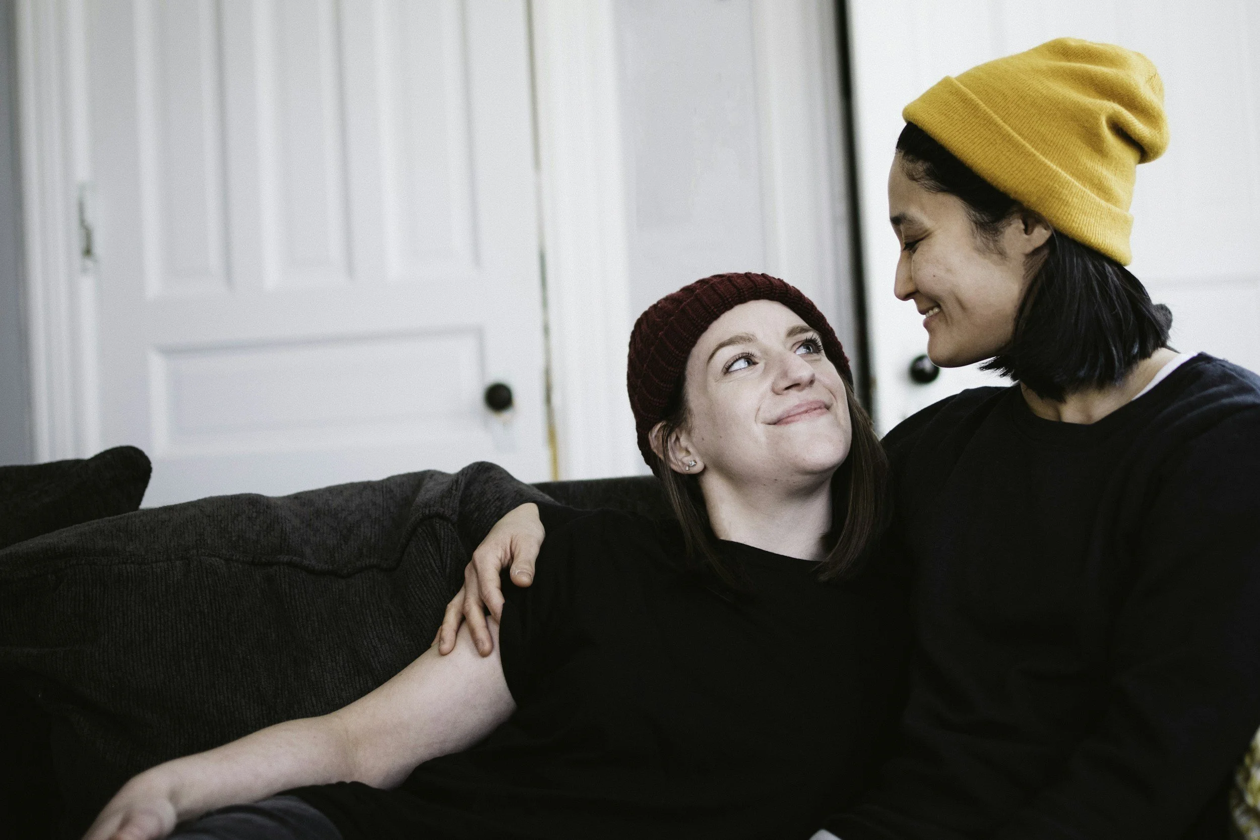 Two women sitting on a black couch, smiling and looking at each other. The woman on the left has light skin, brown hair, and is wearing a red knit beanie and black shirt. The woman on the right has light skin, black hair, and is wearing a yellow beanie and black shirt. The background has white door panels.