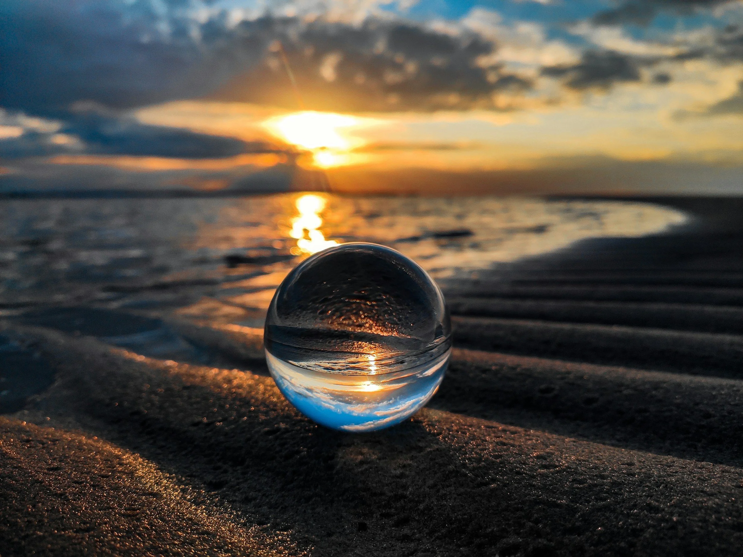 A clear glass sphere on a beach, reflecting the sunset sky and water, with the sun partially obscured by clouds and beautiful orange, yellow, and blue hues.