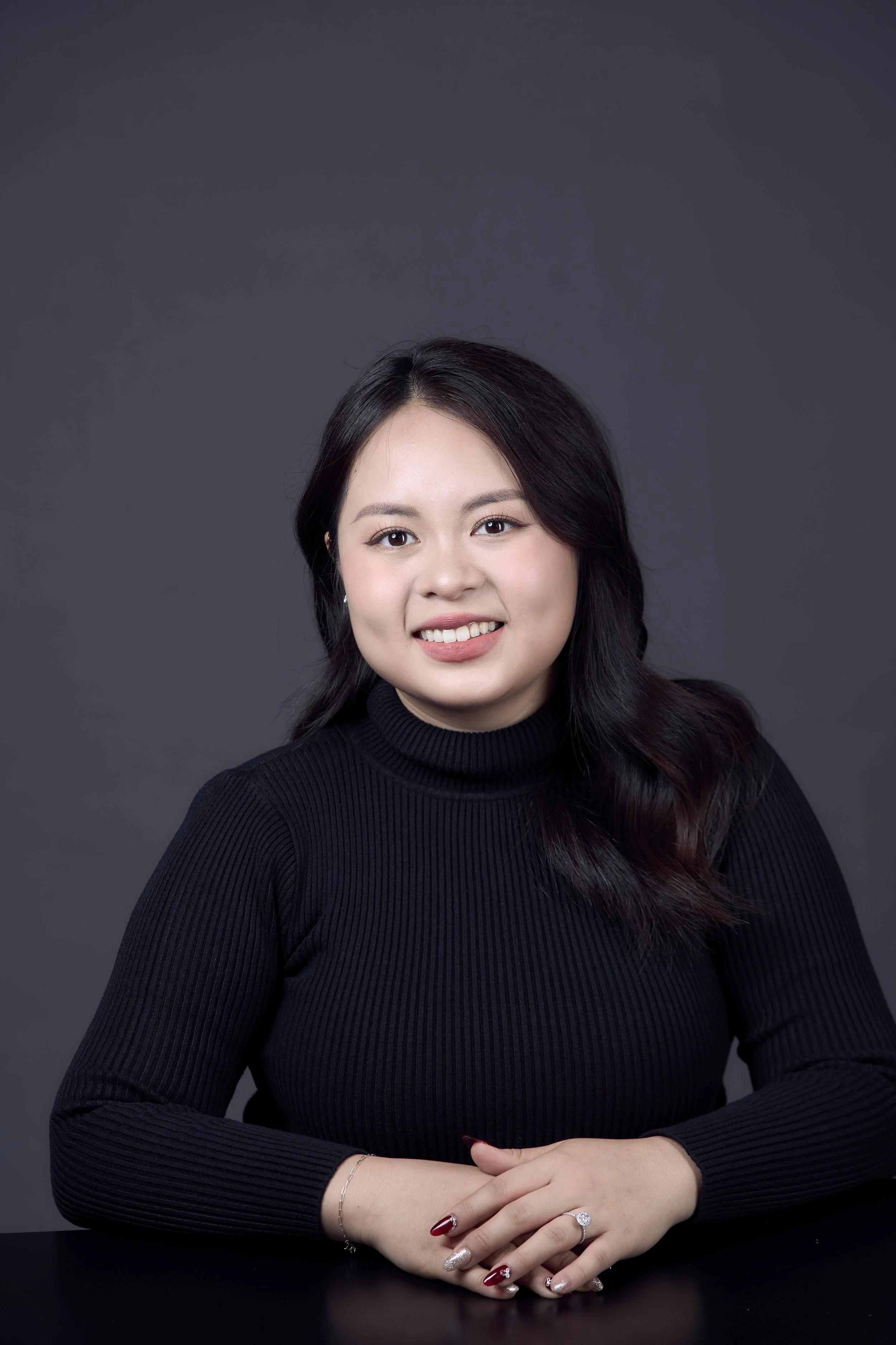A young woman with long dark hair smiling while wearing a black long-sleeve turtleneck sweater, seated at a black table against a dark gray background.