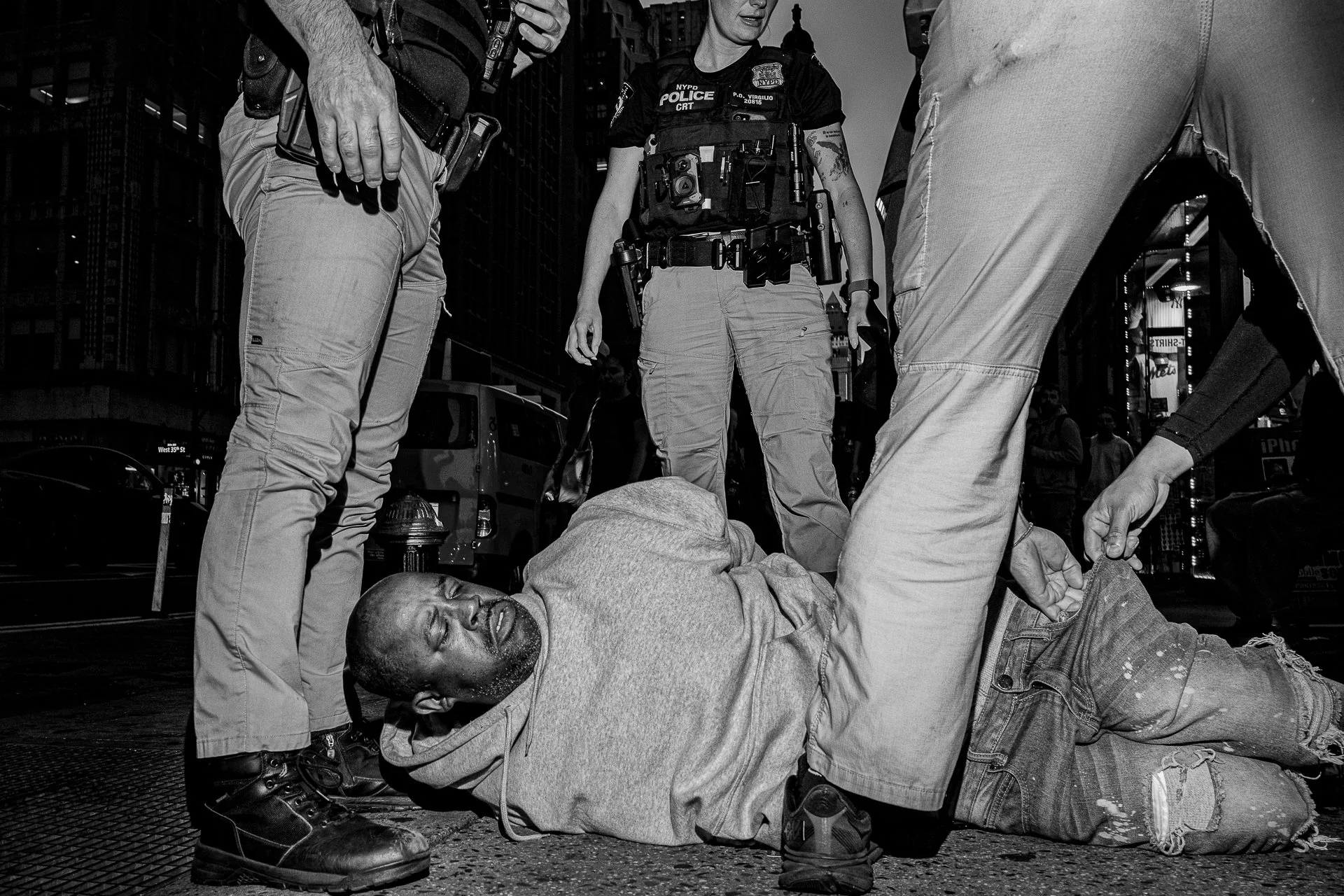 Three police officers surround and assist a kneeling man lying on the ground, in an urban setting at night, with a crowd and city buildings in the background.