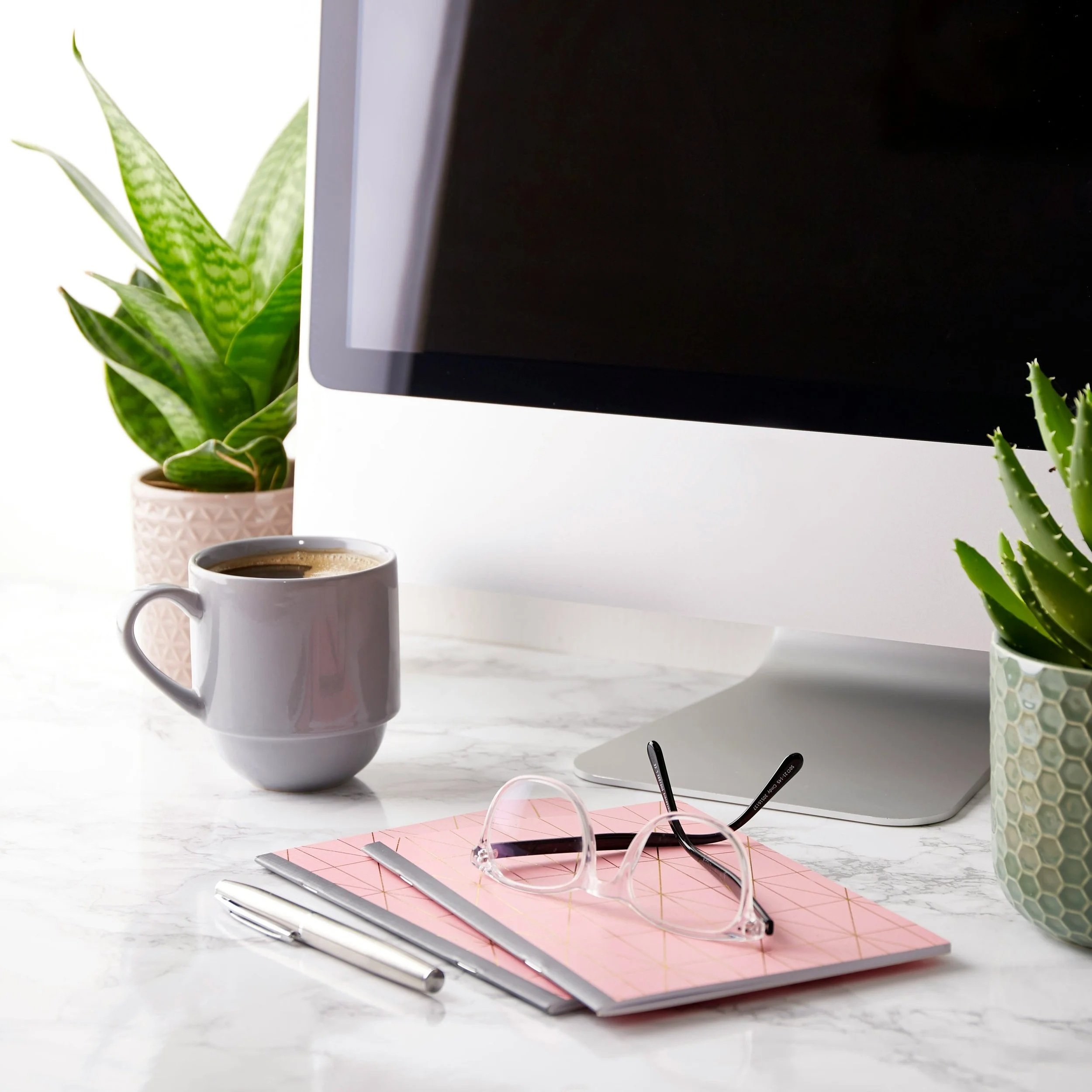 Close-up of a white desk with a cup of coffee, pink glasses, a pink notebook, a pen, and a laptop on a marble surface, with potted plants in the background.