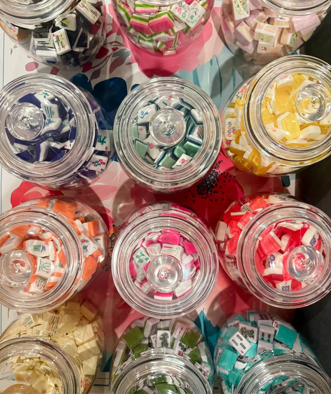 Multiple glass jars filled with colorful Mahjong game tiles in various colors including black, green, yellow, orange, pink, red, cream, and teal, arranged on a table.