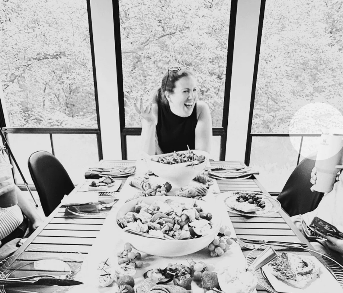 OBB owner Debra laughing at a dining table filled with food, with large windows and trees in the background.