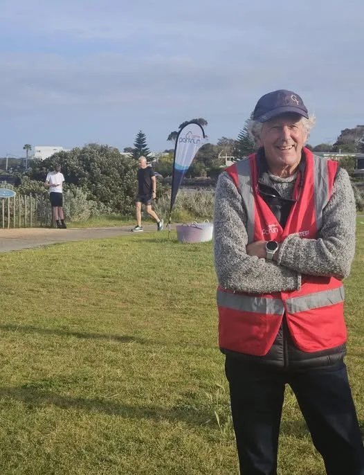 An elderly woman smiling with crossed arms, wearing a gray sweater, red vest, and a navy blue baseball cap, stands on a grassy area outdoors during daytime. In the background, there are three people, one of whom is walking, and two standing near a tr