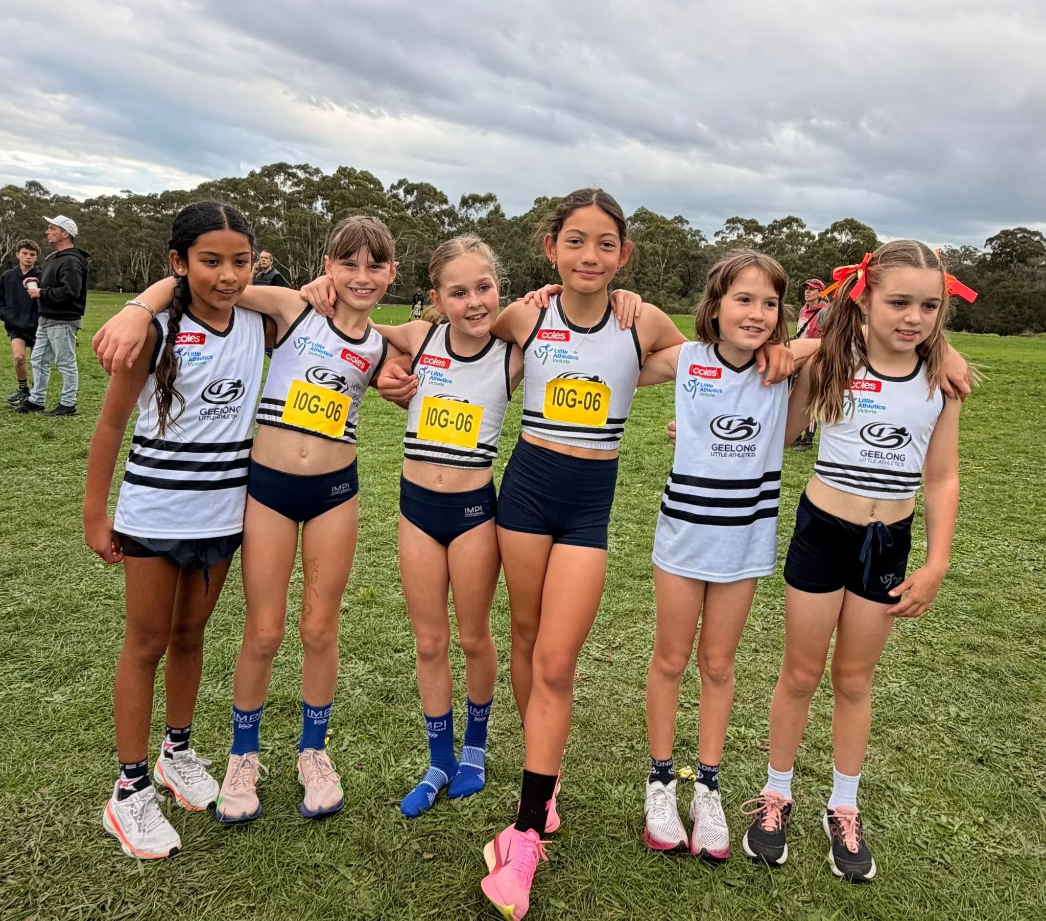A group of six young girls in athletic uniforms standing close together on a grassy field, hugging each other and smiling, with a cloudy sky and other people in the background.