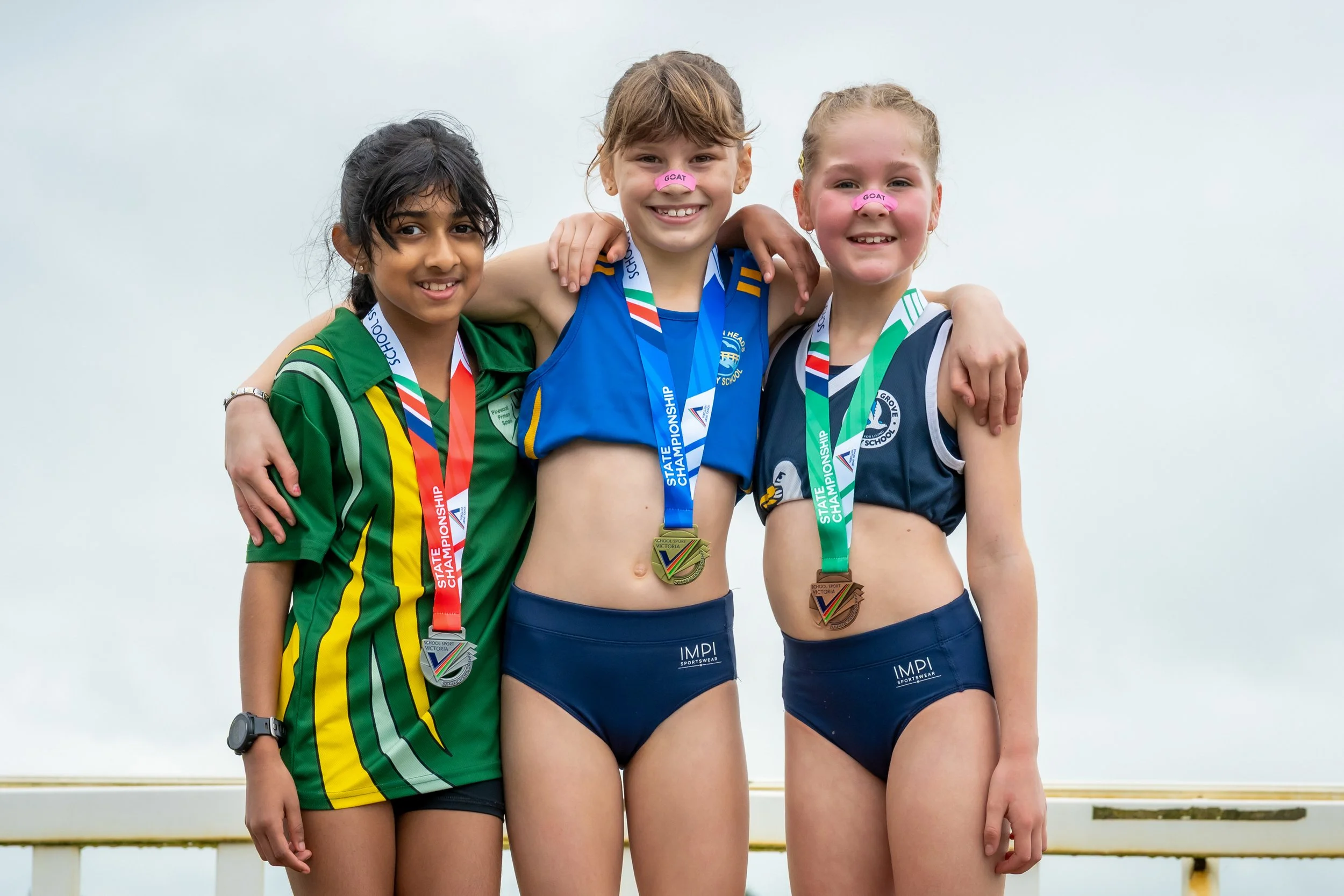 Three young female athletes with medals around their necks pose with arms around each other on a sports field, smiling after a competition.