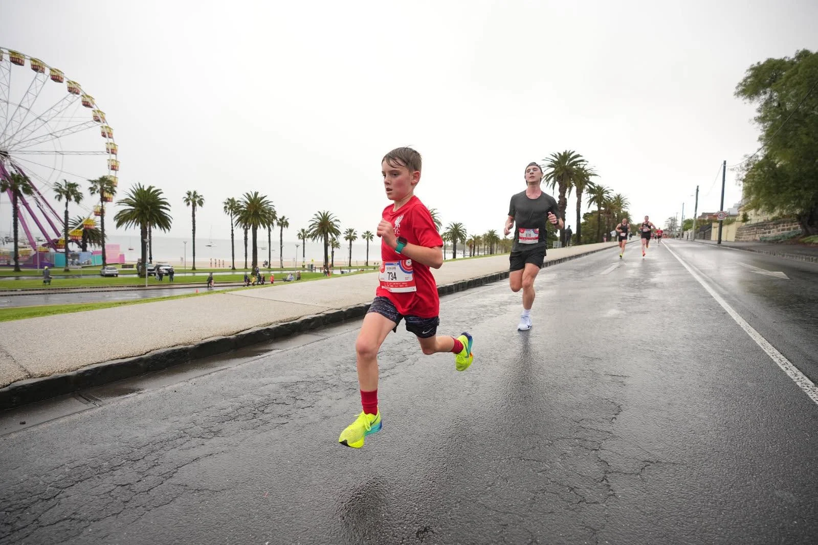 Young boy in red running shirt and shorts participating in a race along a seaside promenade, with several adult runners behind him, nearby palm trees, and a Ferris wheel in the background on a cloudy day.