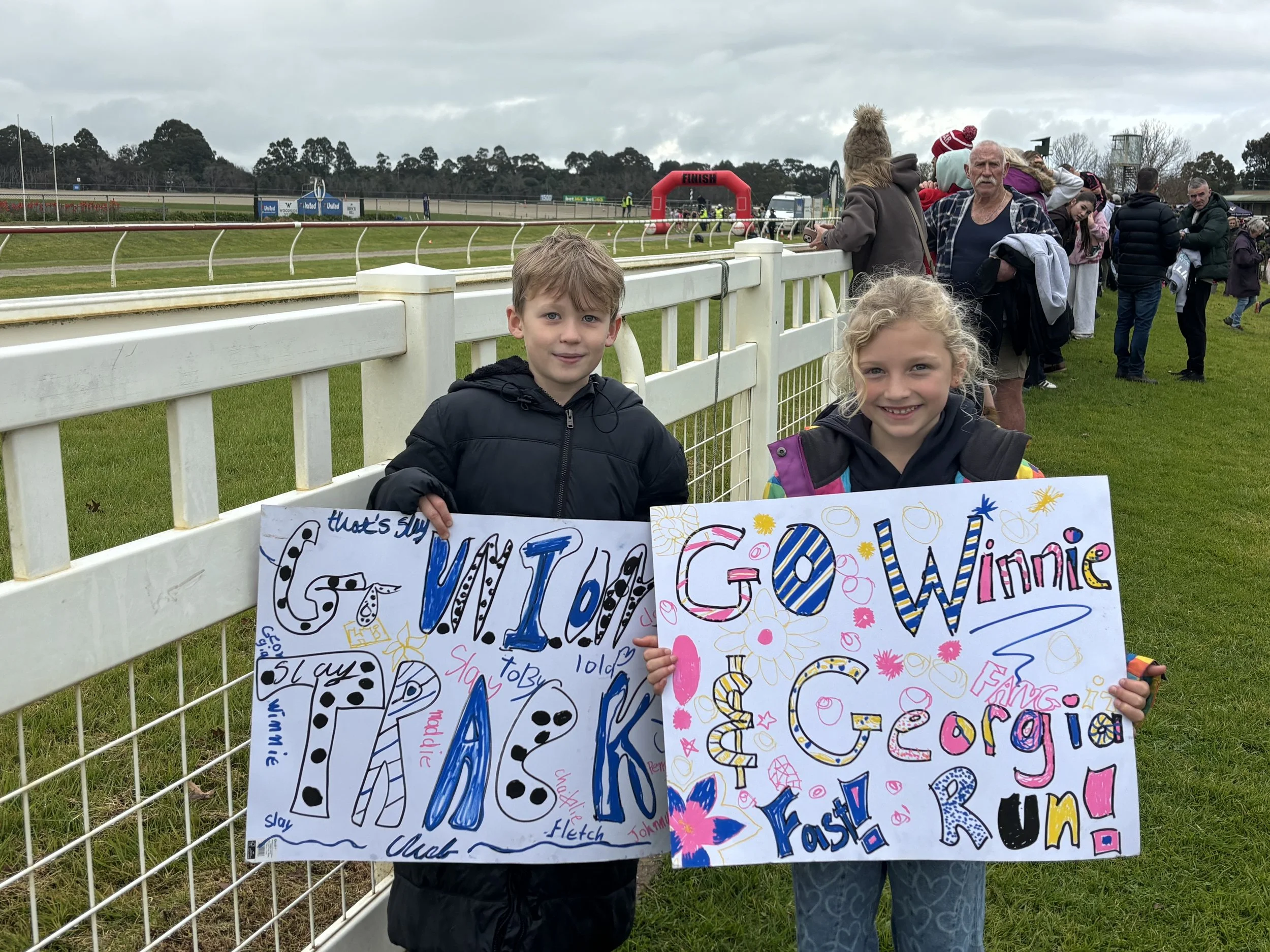Two children standing behind a white fence holding colorful posters with the words "GOAL" and other text, at a race track event with a finish line and spectators in the background.