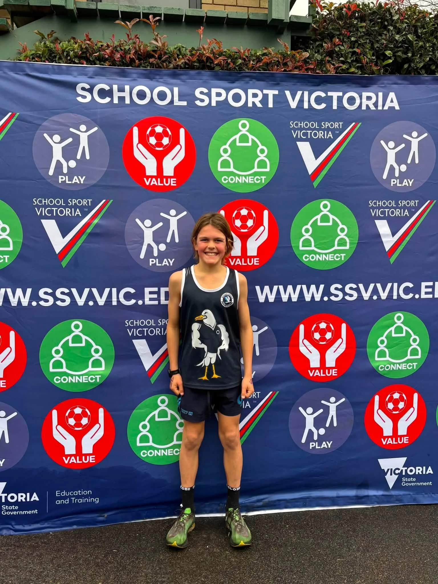 A young girl standing in front of a blue banner with the words 'School Sport Victoria' and multiple icons related to sports and social values, including 'Play', 'Value', and 'Connect'. She is smiling and wearing a sports uniform with a penguin logo, 