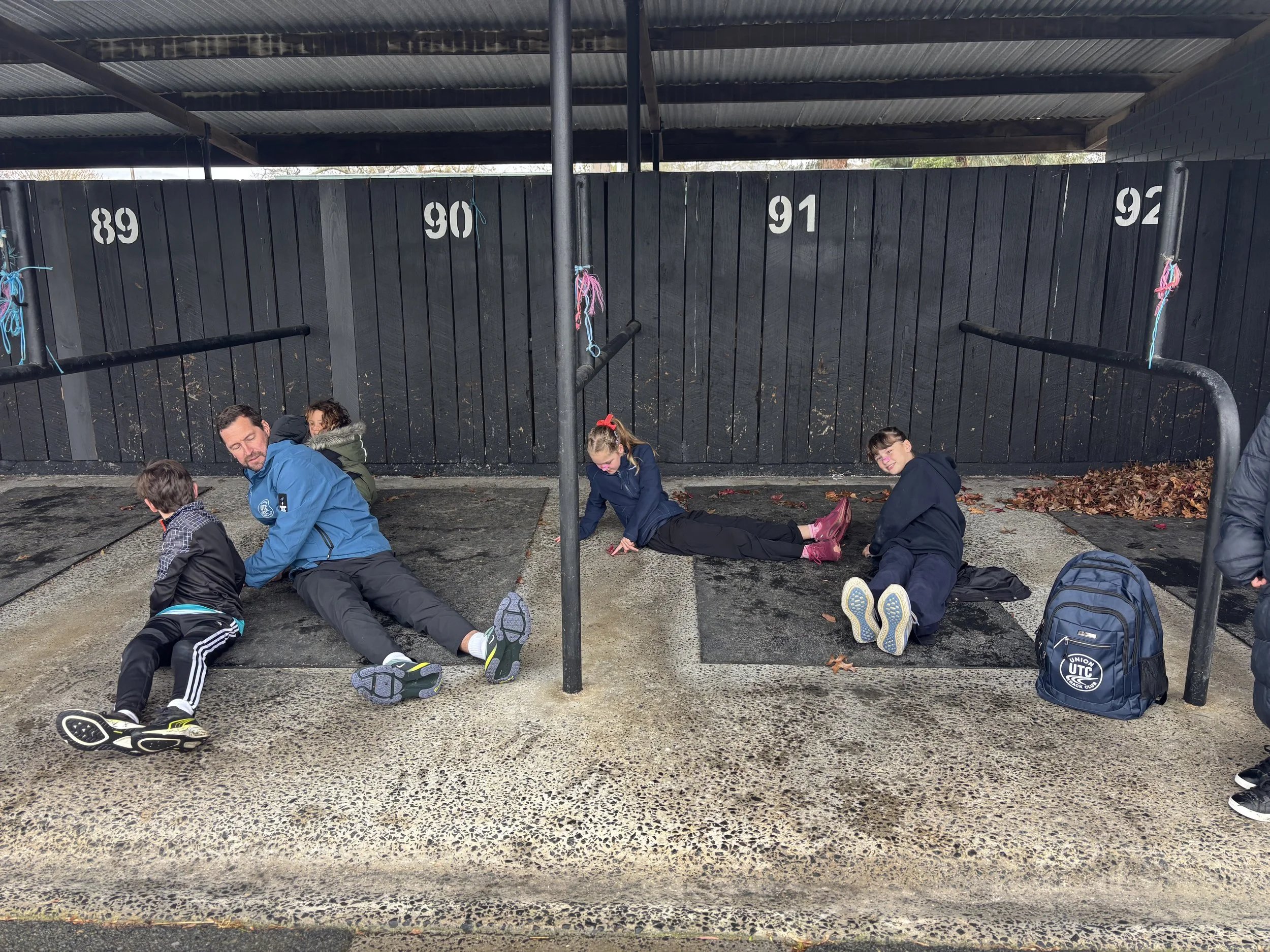 Several children and an adult sitting and lying under a shelter with a black wooden wall numbered 89 to 92 behind them. The children are wearing jackets, and some have backpacks nearby. The ground is paved with leaves scattered around.