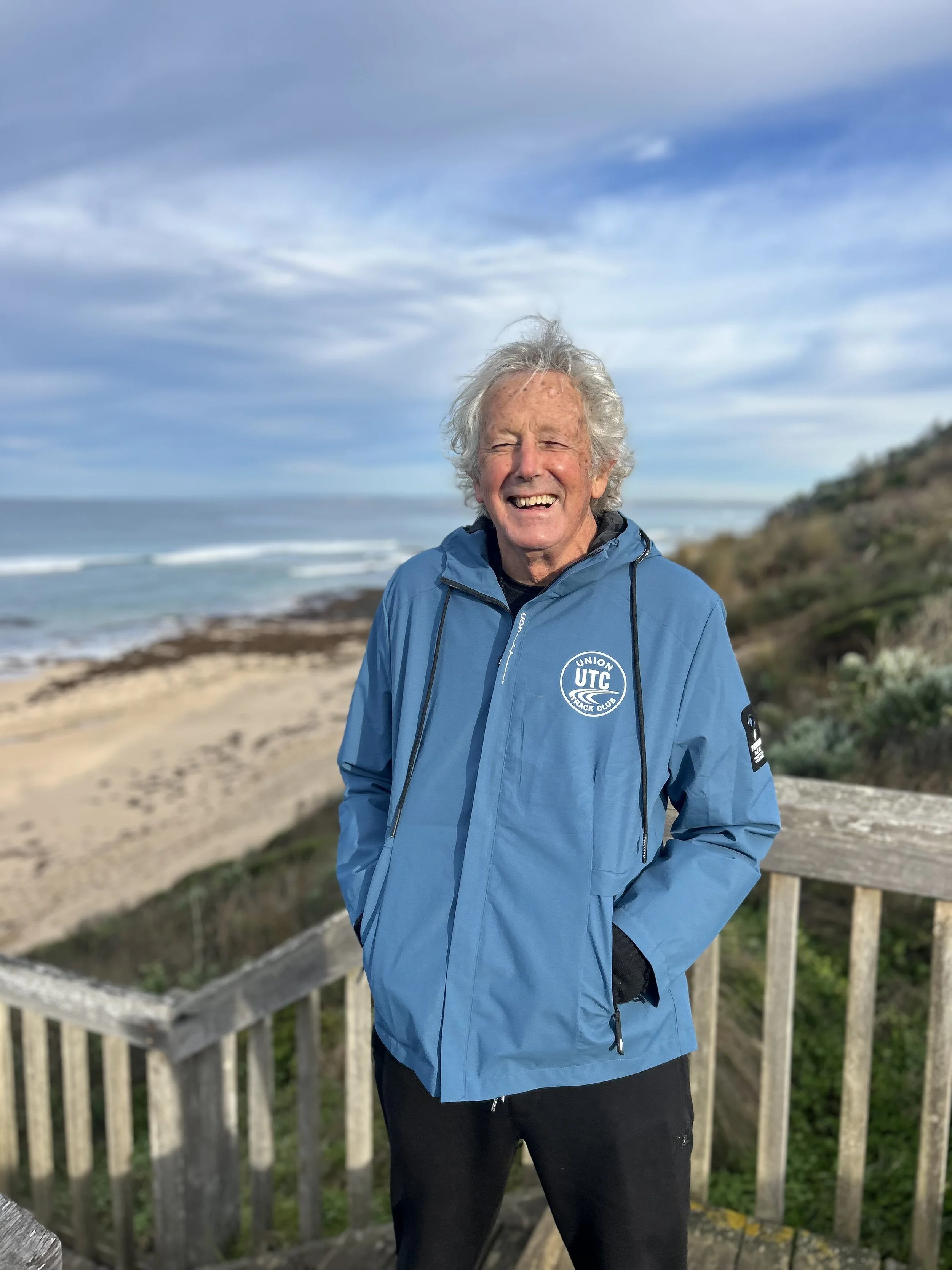 An elderly man with curly white hair smiling outdoors in front of a seascape, wearing a blue jacket with a logo that says "Union UTC Track Club" on it, standing on a wooden platform with a railing, with the ocean and cloudy sky in the background.