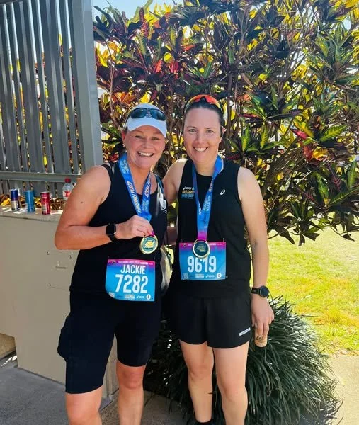 Two women in athletic clothing and medals around their necks, standing outdoors near a hedge, smiling at the camera after a race.