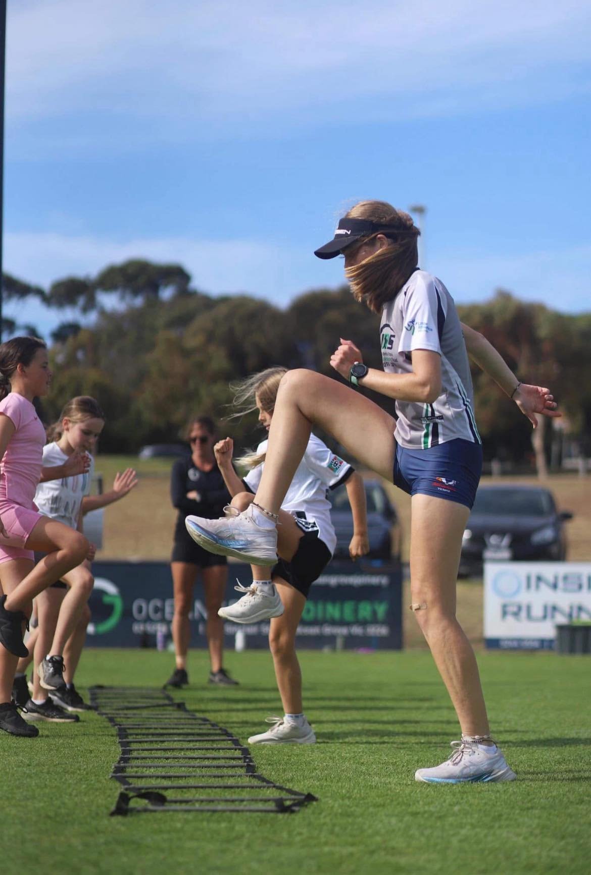 Women participating in a group training session on artificial agility mats outdoors, with a blue sky and trees in the background.