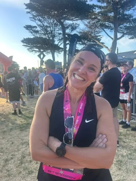 Smiling woman with a medal around her neck, wearing athletic gear, in a park with trees and a crowd of people after running an event.