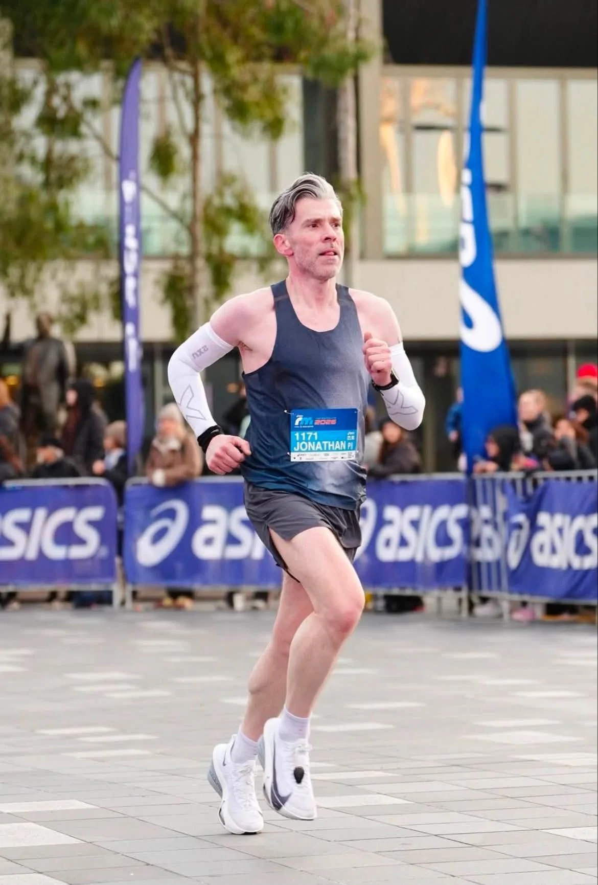 A male runner wearing a blue tank top, black shorts, white arm sleeves, and white running shoes participates in a race. His bib reads 'Jonathan' with the number 1171. He is running on a paved street with a crowd of spectators behind blue barriers and