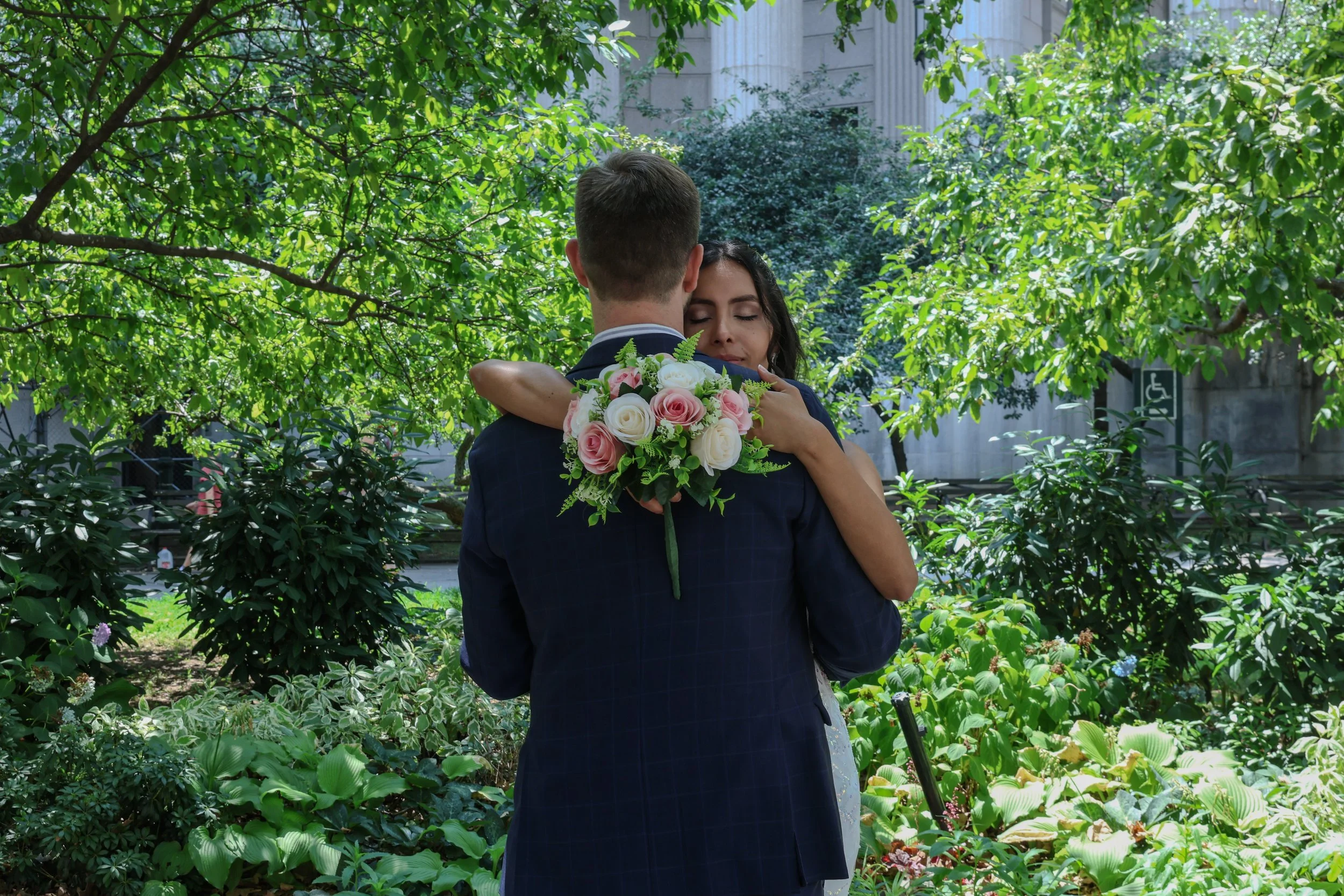 A man and woman embrace in a lush green garden, with the woman holding a bouquet of pink and white roses.