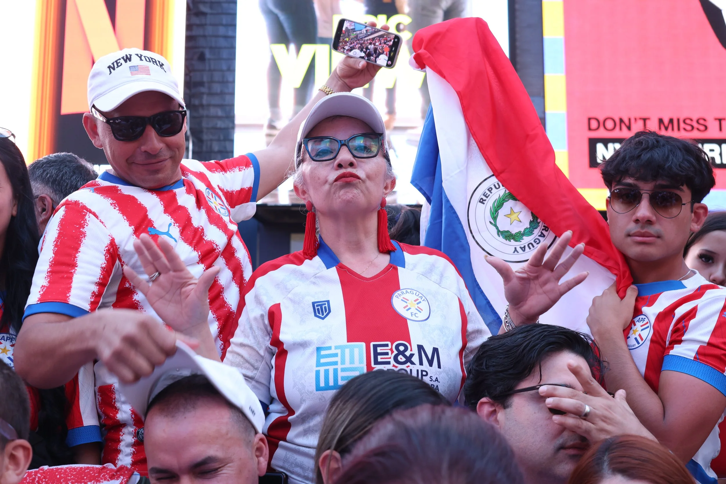 Crowd of people wearing red and white sports jerseys, some wearing sunglasses, with a woman in the center holding a Paraguayan flag, taking a selfie at a sports event.