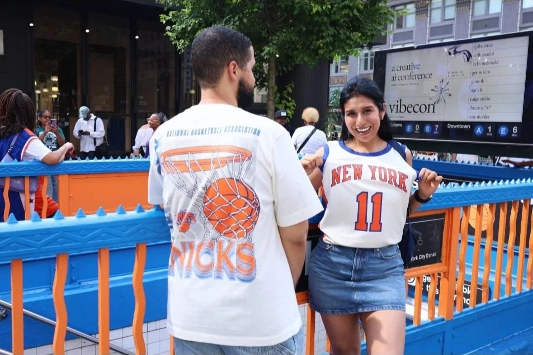 A young woman in a New York Knicks jersey smiling and lifting it with her hands, standing outdoors near a blue railing with a man in a Knicks T-shirt facing her. There are people in the background, some taking pictures, and a digital screen displayin