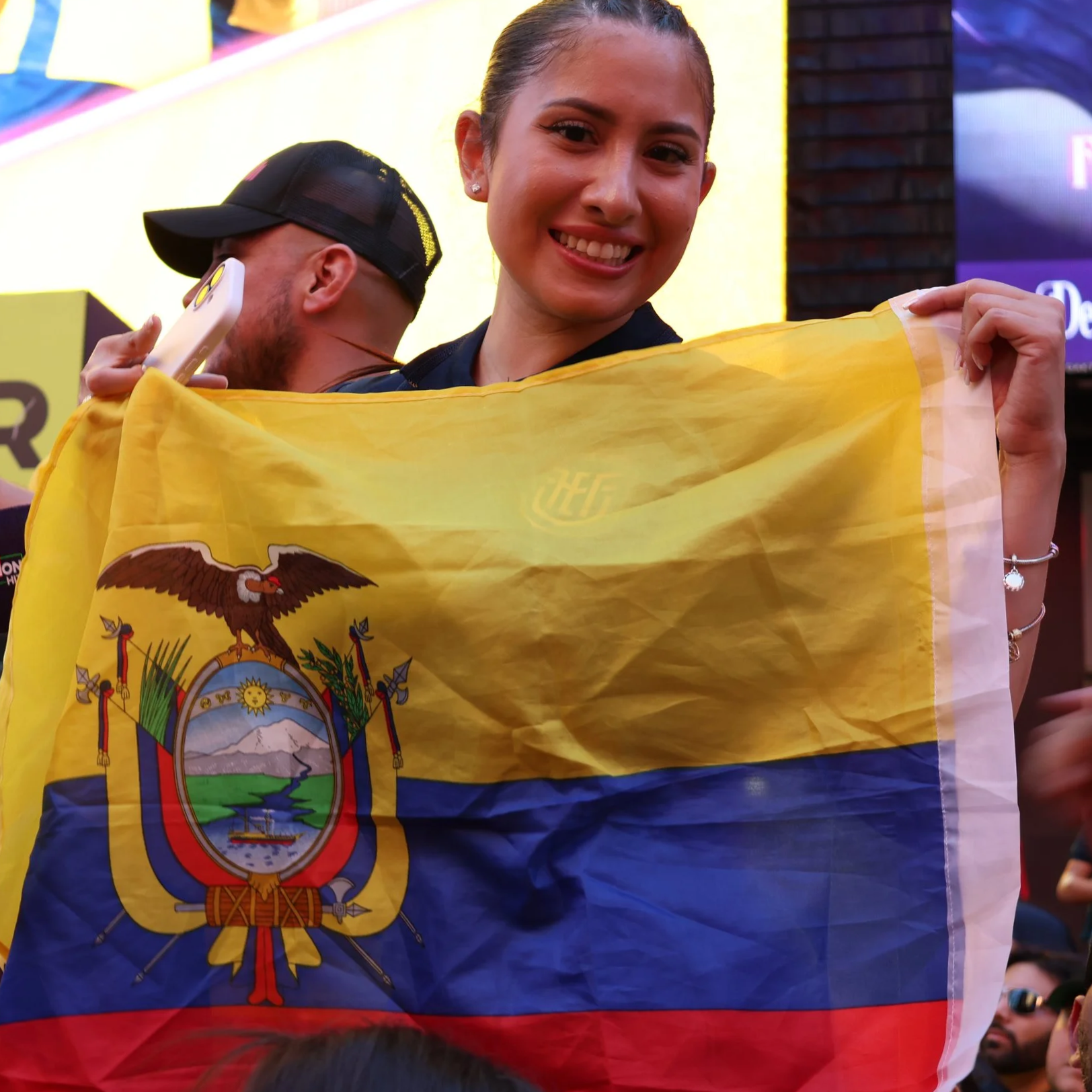 A woman smiling, holding a yellow and blue flag of Ecuador with its coat of arms, at an indoor event.