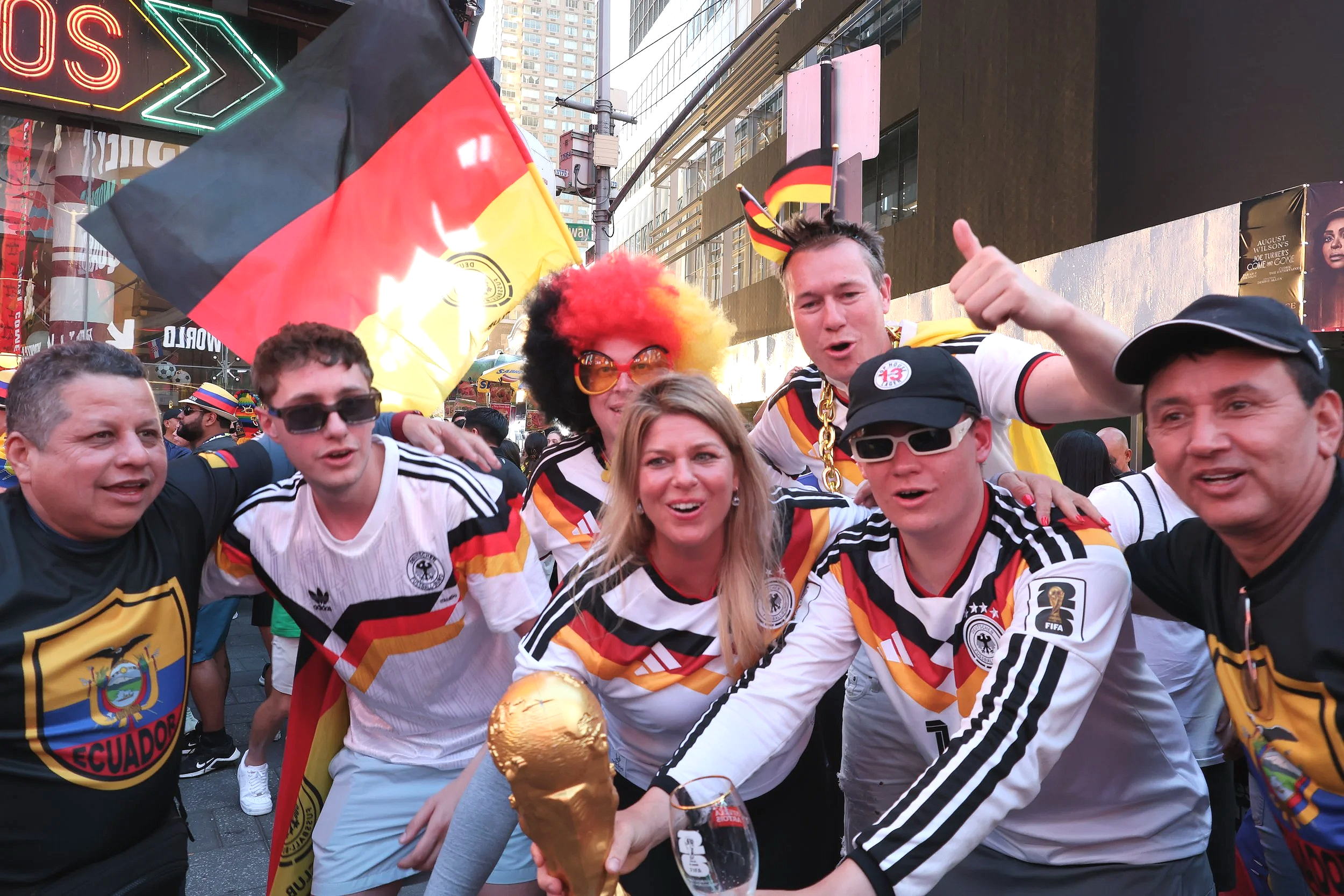 A group of people celebrating, wearing Germany national football team jerseys, some with face paint and accessories in Germany colors, holding a gold trophy, with flags and city buildings in the background.