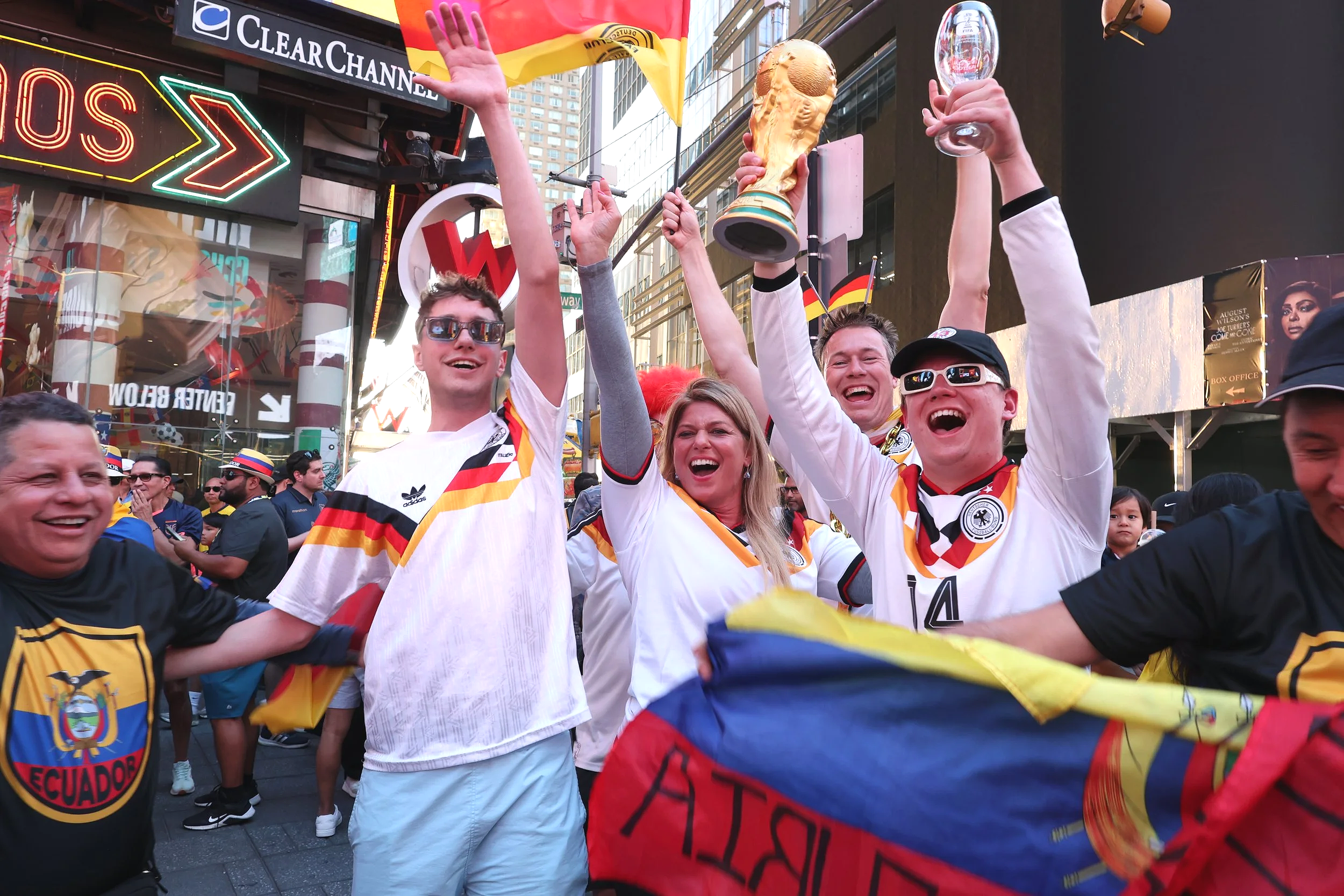 Group of cheering people celebrating on a city street, wearing sports jerseys and waving flags, with some holding a large trophy and others raising their hands.