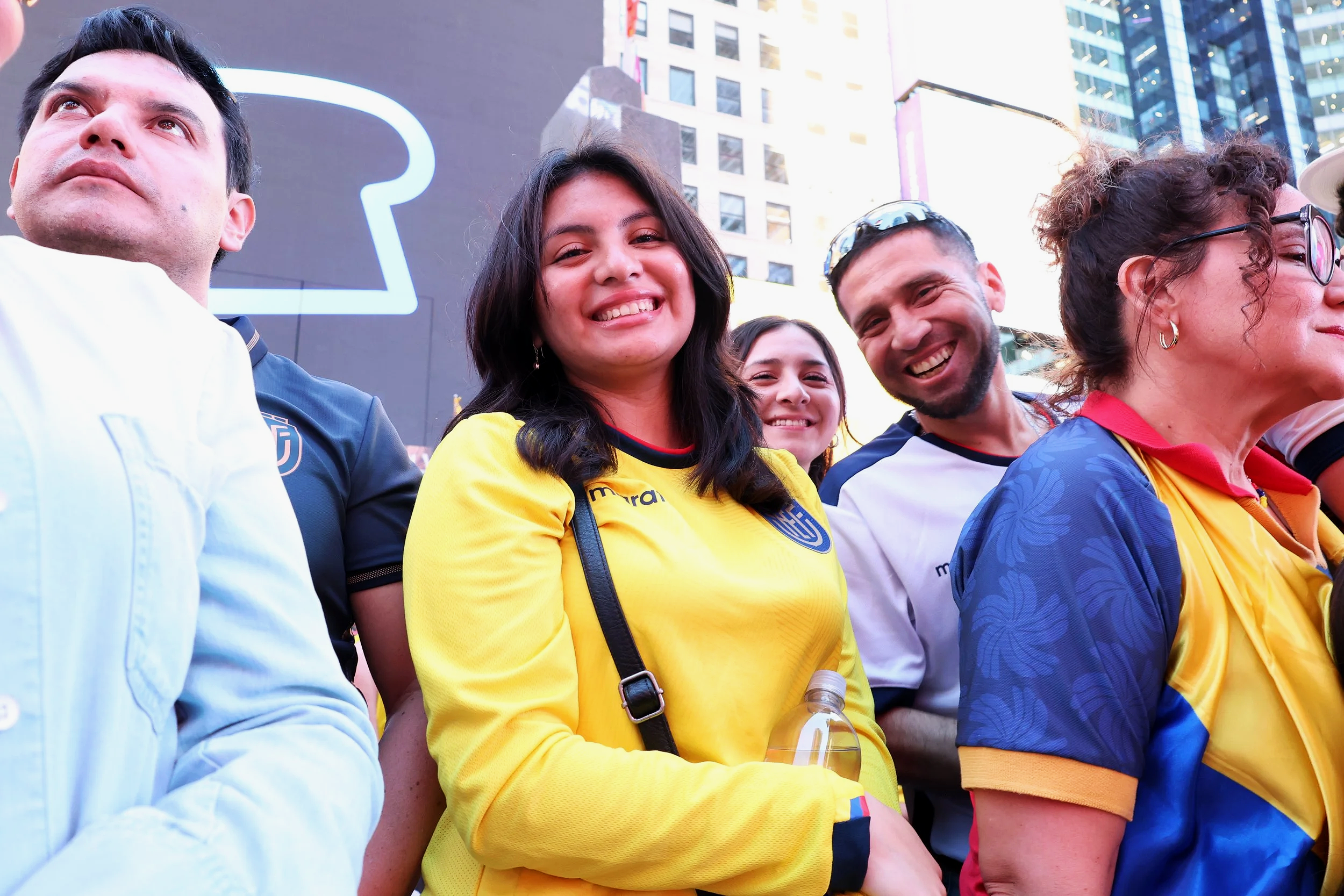 A group of diverse people, including women and men, smiling and standing outdoors in front of tall buildings, some wearing colorful sports jerseys and casual clothing.