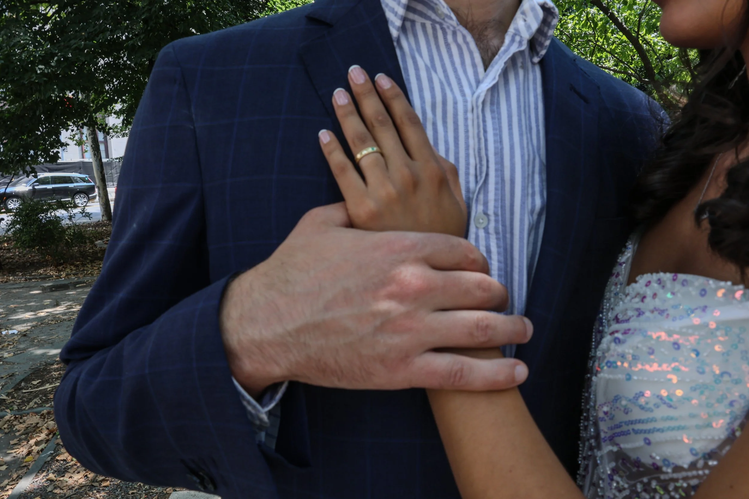 Close-up of a man and woman embracing each other outdoors, with the woman's hand resting on the man's shoulder, displaying a wedding ring.