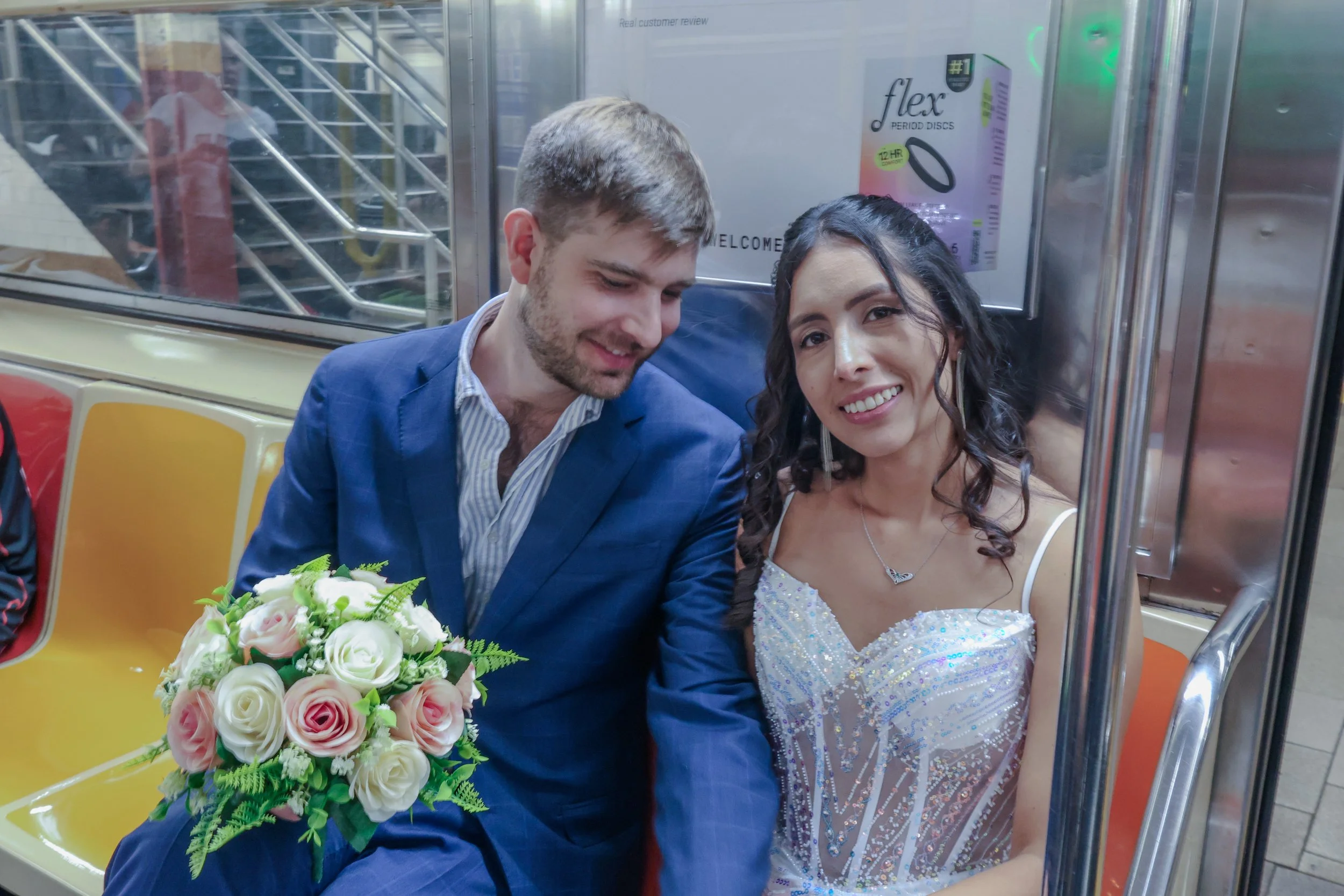 A couple dressed in wedding attire sitting together in a subway train, with the woman holding a bouquet of pink and white roses. The man is wearing a blue suit and the woman is wearing a white dress with silver embellishments, smiling at the camera.