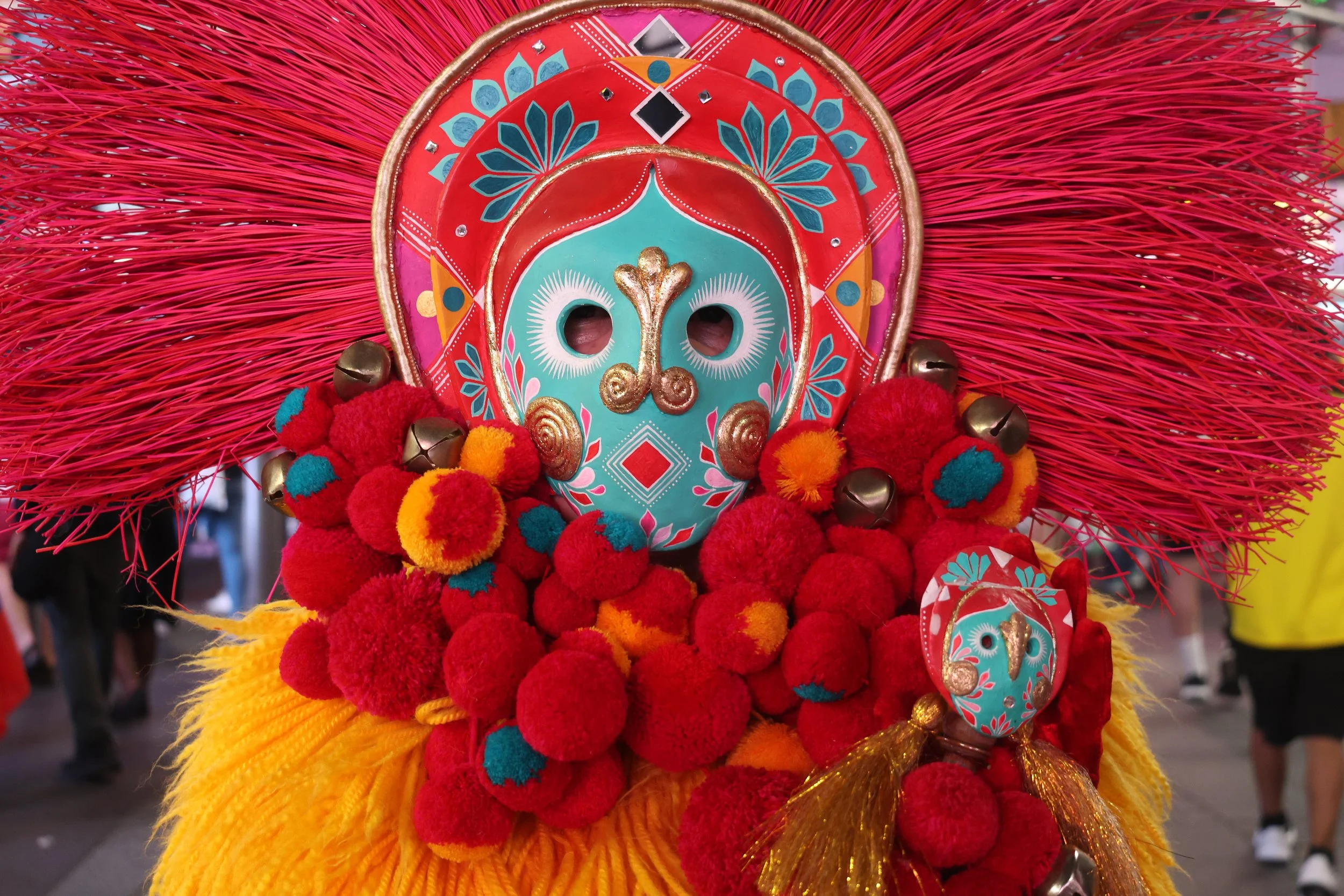 Colorful traditional costume with a decorated mask, surrounded by pom-poms and cloth, likely worn in cultural festival or celebration.