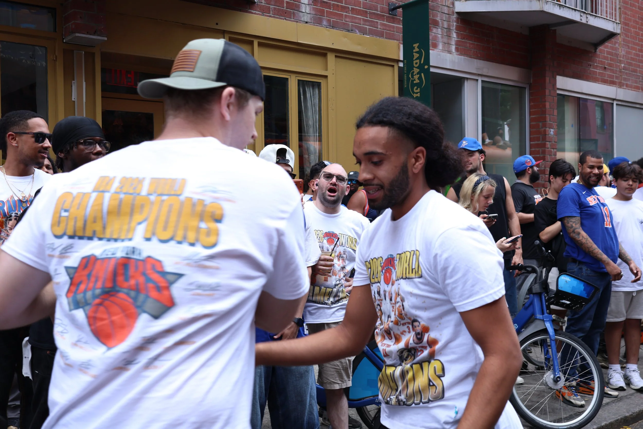 A group of people gathered outdoors, with some wearing New York Knicks World Champions T-shirts, chatting and socializing near a brick building on a city street.